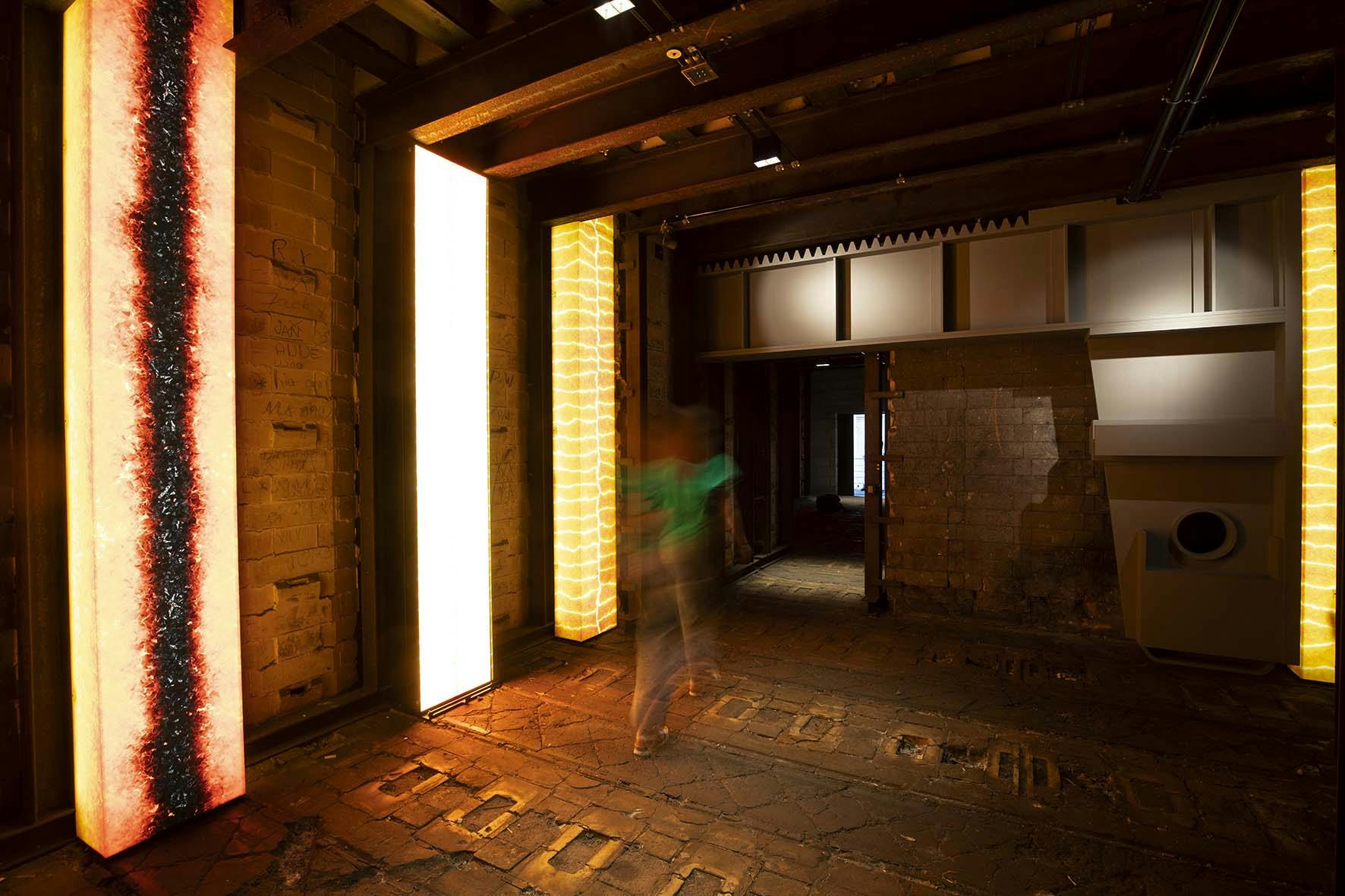Ein dunkler Raum mit industriellem Charakter wird von vertikalen, leuchtenden Lichtinstallationen in warmen Farben erhellt. Die Wände bestehen aus alten Ziegeln, der Boden ist uneben und aus Steinplatten. Eine verschwommene Person bewegt sich durch den Raum, was auf eine Langzeitbelichtung hinweist. Im Hintergrund führt ein Durchgang in einen weiteren abgedunkelten Raum.