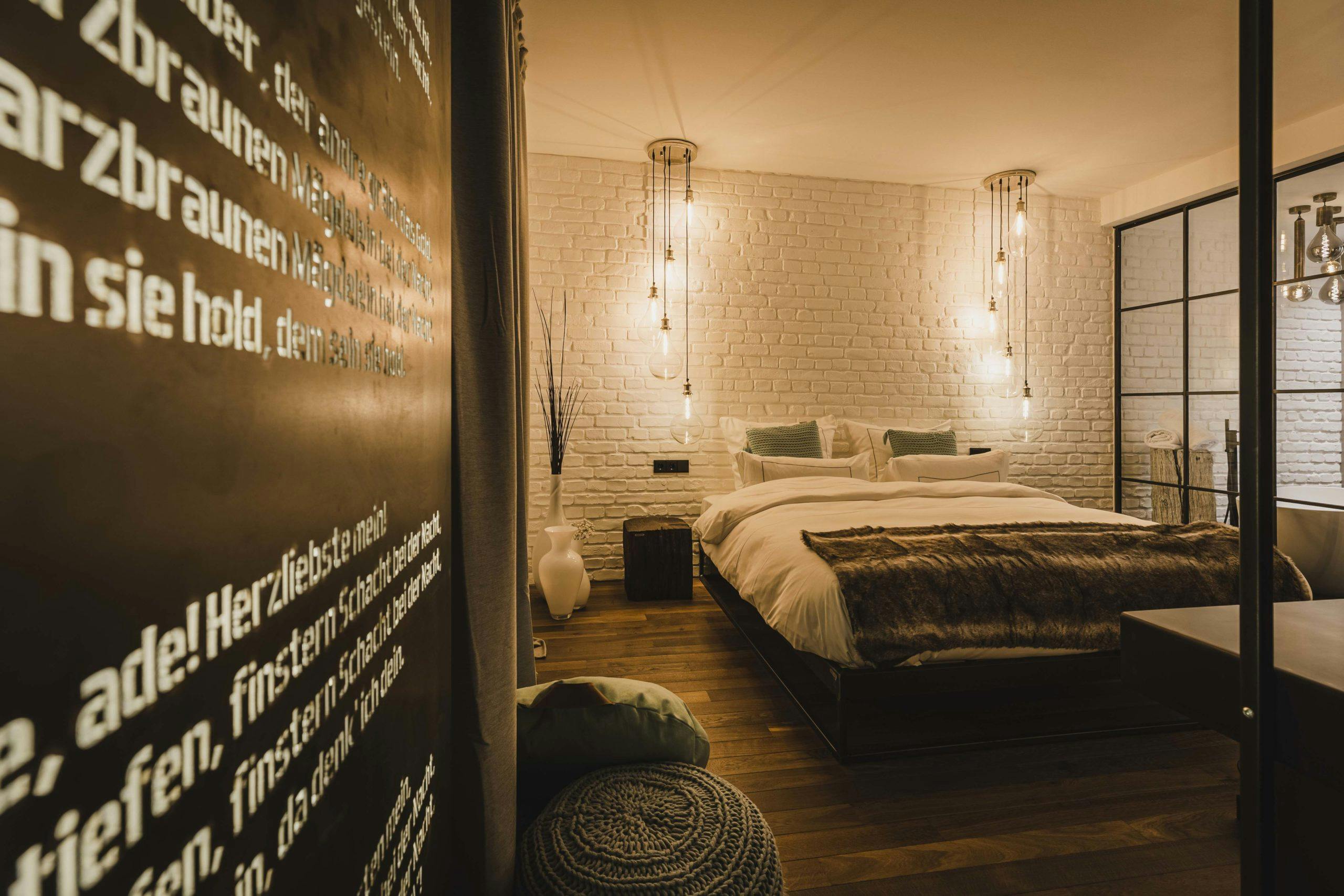 Stilvoll eingerichtetes Schlafzimmer mit einem großen Doppelbett, weißen Kissen und Decke sowie einem braunen Fellüberwurf am Fußende. Die Rückwand besteht aus weiß gestrichenem Backstein, davor hängen mehrere dekorative Glühbirnen an langen Kabeln von der Decke. Links im Bild ist eine schwarze Wandtafel mit Auszügen aus dem Steigerlied in weißer Frakturschrift zu sehen. Der Raum ist in warmes Licht getaucht, auf dem dunklen Holzboden liegen ein graues Strickkissen und ein helles Sitzkissen. Im Hintergrund ist ein angrenzendes Badezimmer mit freistehender Badewanne sichtbar.