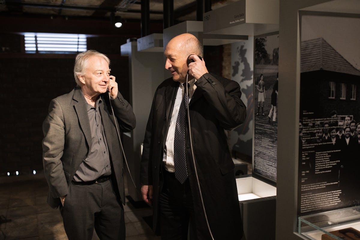 Zwei ältere Männer in Anzügen stehen nebeneinander in einer Ausstellung und halten jeweils einen Telefonhörer an ihr Ohr. Sie wirken interessiert und lächeln einander an, als würden sie eine Audiostation gemeinsam erleben. Im Hintergrund sind beleuchtete Informationstafeln mit historischen Fotos und Texten zu sehen. Die Szene spielt in einem abgedunkelten Raum mit musealem Charakter.