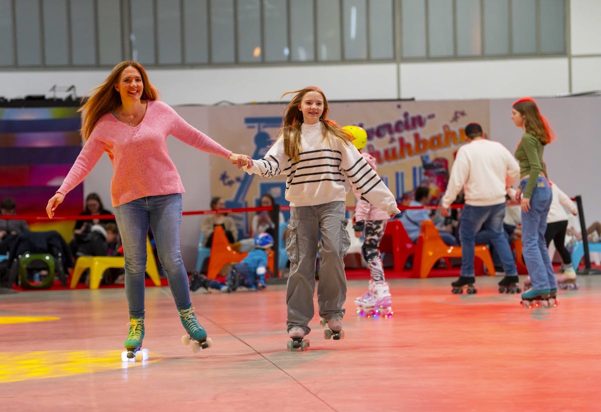 In einer bunten Rollschuhhalle fahren eine Frau in rosa Pullover und ein Mädchen mit gestreiftem Pullover und grauer Hose Hand in Hand auf Rollschuhen über die Bahn. Beide lachen und wirken fröhlich. Im Hintergrund fahren weitere Personen auf Rollschuhen, während einige Menschen auf bunten Sitzgelegenheiten am Rand der Halle sitzen. Die Atmosphäre ist lebendig und freizeitbetont.