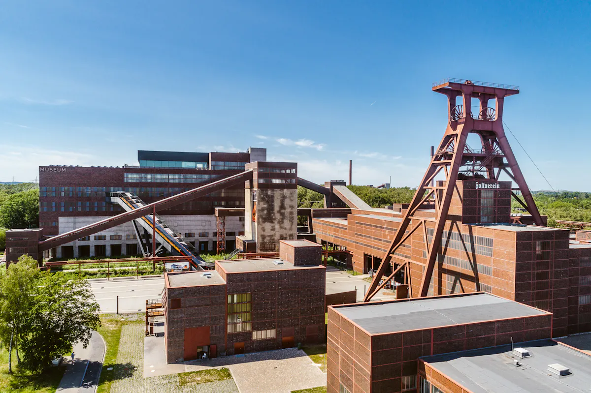 Luftaufnahme des UNESCO-Welterbes Zollverein in Essen bei sonnigem Wetter. Im Vordergrund ist das markante Fördergerüst der Zeche mit der Aufschrift Zollverein zu sehen. Das Gelände besteht aus roten Backsteingebäuden mit großen Fensterflächen und wird von schrägen Förderbändern durchzogen. Links im Bild ist das Ruhr Museum erkennbar. Im Hintergrund erstreckt sich eine grüne Baumlandschaft. Der Himmel ist klar und blau.