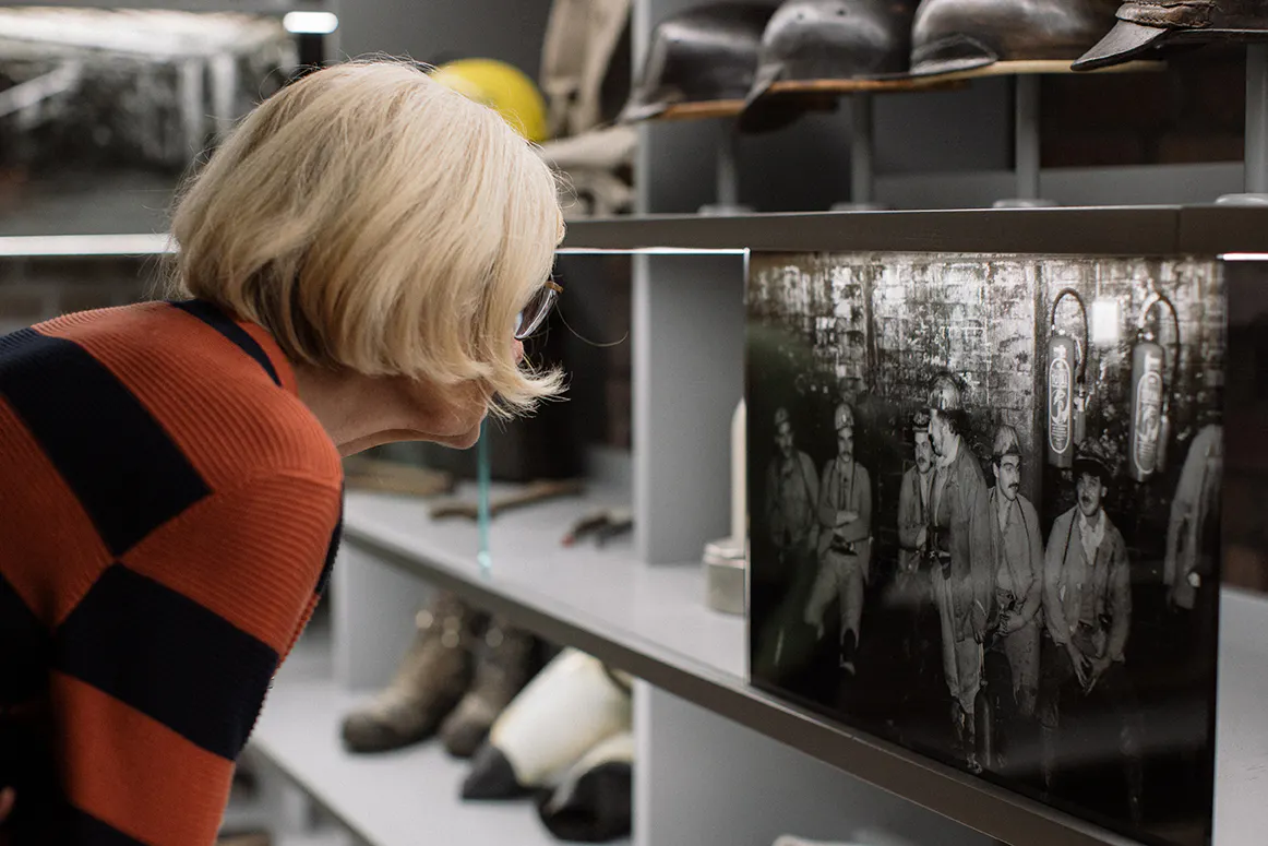 Eine ältere Frau mit blonden Haaren und Brille betrachtet aufmerksam ein historisches Schwarz-Weiß-Foto, das in einer Museumsvitrine ausgestellt ist. Das Foto zeigt eine Gruppe von Bergleuten in Arbeitskleidung mit Helmen, die offenbar in einem untertägigen Aufenthaltsraum sitzen oder stehen. Um die Vitrine herum sind weitere Ausstellungsstücke wie alte Helme, Stiefel und Werkzeuge aus dem Bergbau zu sehen. Die Szene spielt sich in einem Raum mit gedämpftem Licht ab, der eine museale Atmosphäre vermittelt.