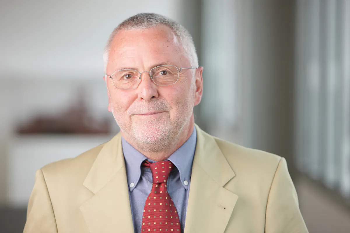 Ein älterer Mann mit kurzem grauem Haar, Brille und gepflegtem Bart steht vor einem unscharfen Hintergrund mit großen Fenstern. Er trägt ein beiges Sakko, ein blaues Hemd und eine rote Krawatte mit kleinen gelben Quadraten und blickt freundlich in die Kamera.
