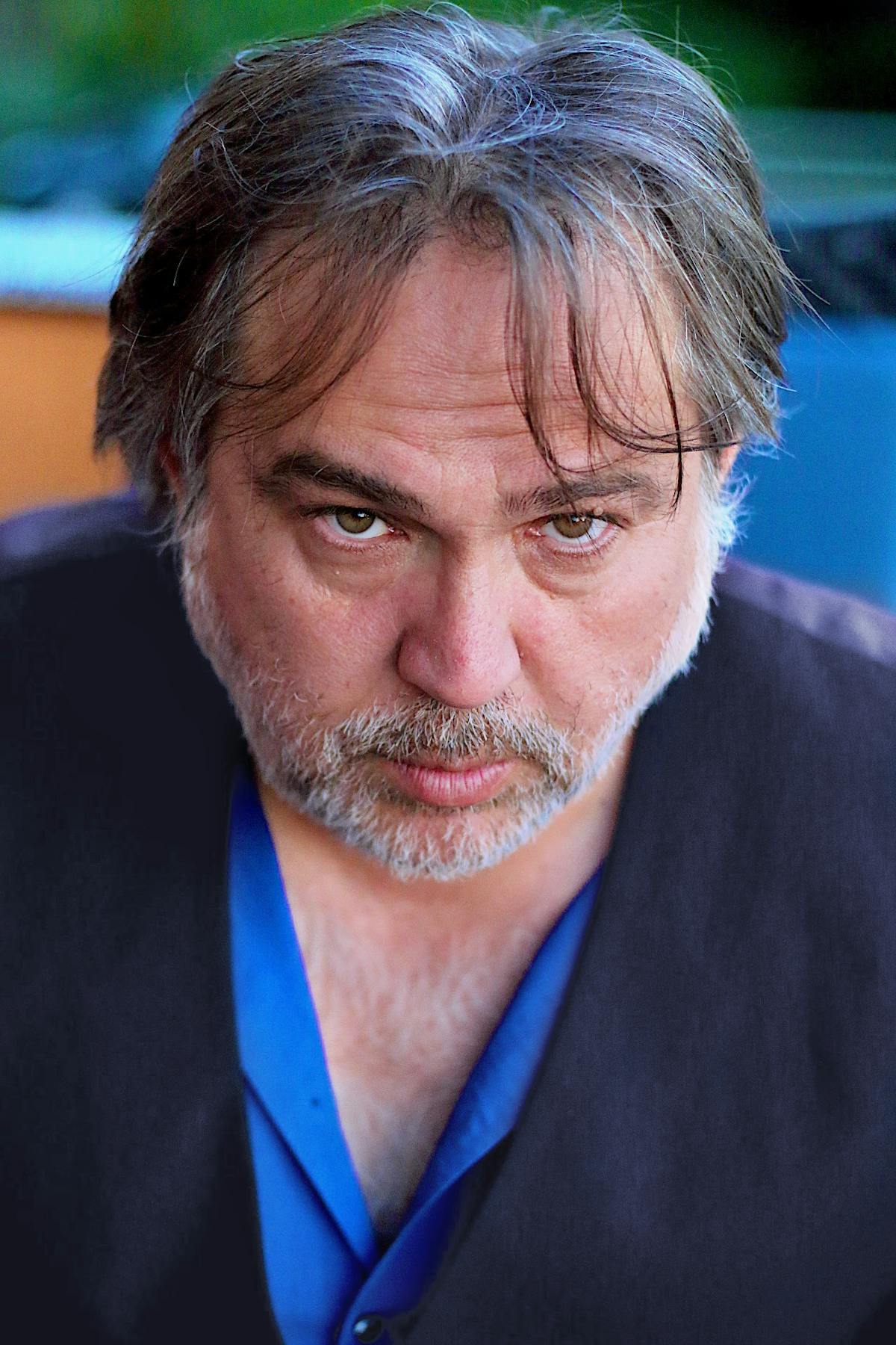 Porträt eines Mannes mittleren Alters mit graumelierten, leicht zerzausten Haaren und Bartstoppeln. Er trägt ein blaues Hemd unter einer dunklen Weste. Der Mann blickt ernst und leicht nach unten gerichtet in die Kamera. Der Hintergrund ist unscharf und zeigt grüne und blaue Farbtöne.