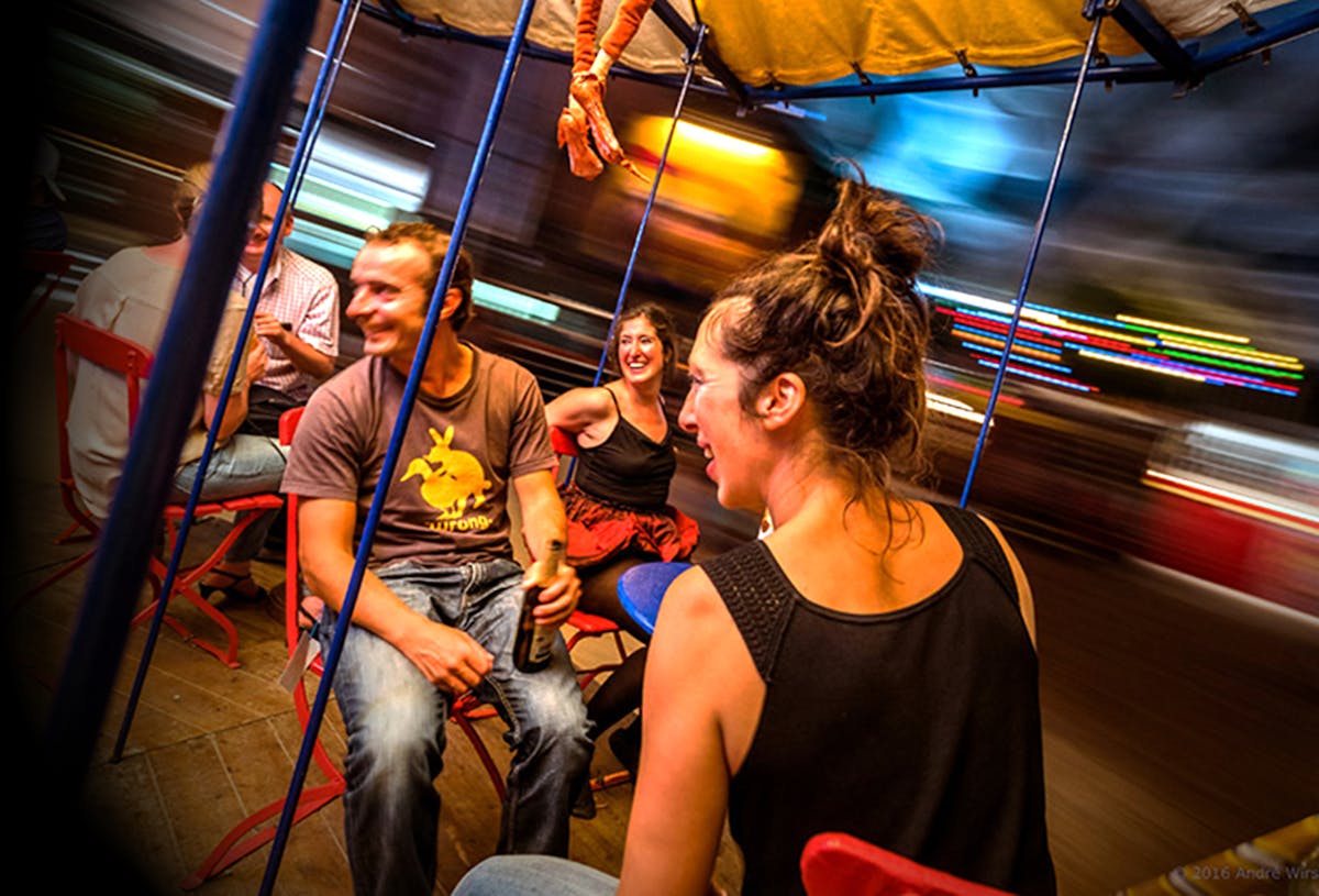 Mehrere Menschen sitzen auf roten Stühlen in einem sich drehenden Karussell mit Zeltdach. Sie lachen, unterhalten sich und wirken ausgelassen. Die Bewegung des Karussells ist durch die verschwommenen Lichter und Hintergrundelemente sichtbar. Ein Mann hält eine Bierflasche in der Hand, eine Frau im Vordergrund lacht herzlich. Die Stimmung ist fröhlich und lebendig, es scheint sich um ein besonderes, interaktives Fahrgeschäft oder Kunstprojekt zu handeln.