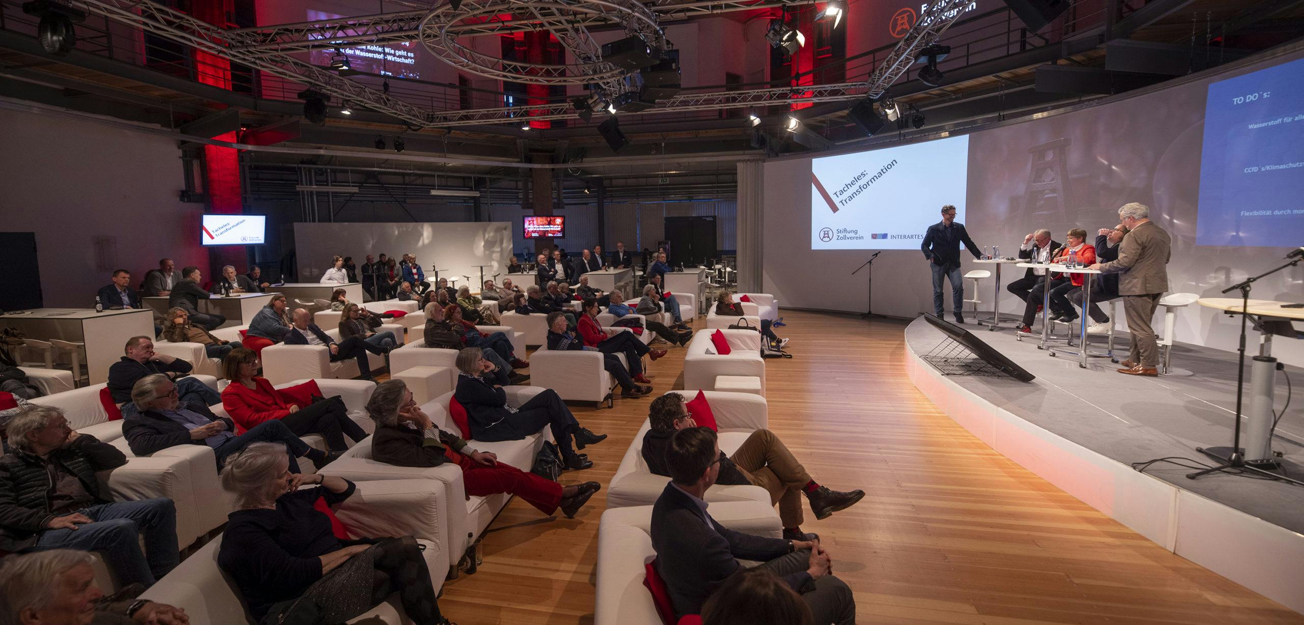Eine große Gruppe von Menschen sitzt auf weißen Sofas in einem modernen Veranstaltungsraum mit Holzboden. Im Vordergrund und seitlich sitzt das Publikum, aufmerksam einer Podiumsdiskussion folgend. Auf der Bühne stehen fünf Personen an einem Stehtisch mit Mikrofonen, links daneben ein Moderator. Im Hintergrund ist eine große Projektionsfläche mit dem Titel der Veranstaltung Tacheles: Transformation sowie Logos der Stiftung Zollverein und Interartes zu sehen. Der Raum ist mit roter Beleuchtung und Technikträgern an der Decke ausgestattet. Die Atmosphäre wirkt professionell und dialogorientiert.