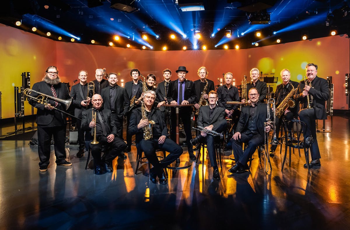 Eine Bigband mit siebzehn Musikerinnen und Musikern steht und sitzt in einem halbrunden Arrangement auf einer Bühne mit gelb-orangefarbener Beleuchtung. Die Bandmitglieder tragen dunkle Kleidung, viele halten Blasinstrumente wie Trompeten, Posaunen und Saxophone in der Hand. In der Mitte steht ein Mann mit schwarzem Hut und violetter Krawatte an einem Stehtisch. Im Hintergrund sorgen Bühnenlichter und blauer Scheinwerfernebel für eine atmosphärische Beleuchtung.