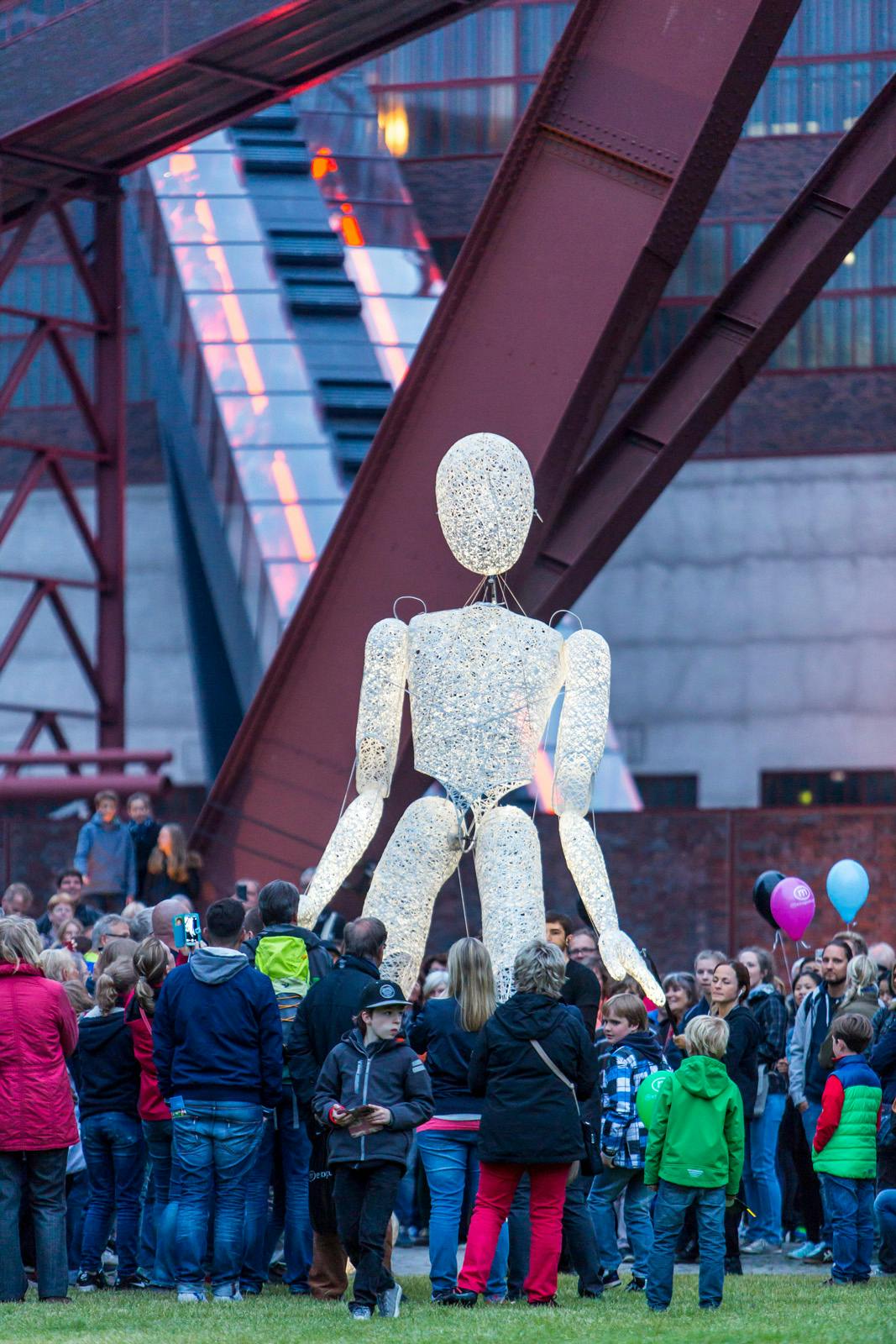 Eine große, beleuchtete Figur in menschlicher Form aus einem durchscheinenden, netzartigen Material steht inmitten einer Menschenmenge auf einer Wiese. Die Figur ist deutlich größer als die umstehenden Personen und wirkt wie eine bewegliche Puppe oder Skulptur. Um sie herum stehen viele Erwachsene und Kinder, einige mit Luftballons, andere fotografieren das Spektakel. Im Hintergrund sind rote Stahlträger und ein mit Licht beleuchtetes Gebäude zu sehen, vermutlich auf dem Gelände des UNESCO-Welterbes Zeche Zollverein. Die Szene wirkt wie ein Teil einer öffentlichen Kunst- oder Kulturveranstaltung.