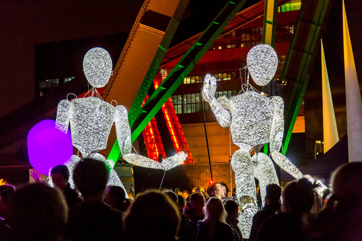 Zwei große, leuchtende Figuren aus einem netzartigen Material stehen auf einem nächtlich beleuchteten Platz, umgeben von vielen Menschen. Die Figuren wirken wie überlebensgroße Marionetten mit menschlicher Gestalt und werden durch Stäbe von Personen geführt. Im Hintergrund sind beleuchtete Industriearchitektur und bunte Lichtinstallationen in grün, rot und orange zu sehen, vermutlich auf dem Gelände der Zeche Zollverein. Die Szene vermittelt den Eindruck einer festlichen Veranstaltung mit Kunst und Performance.