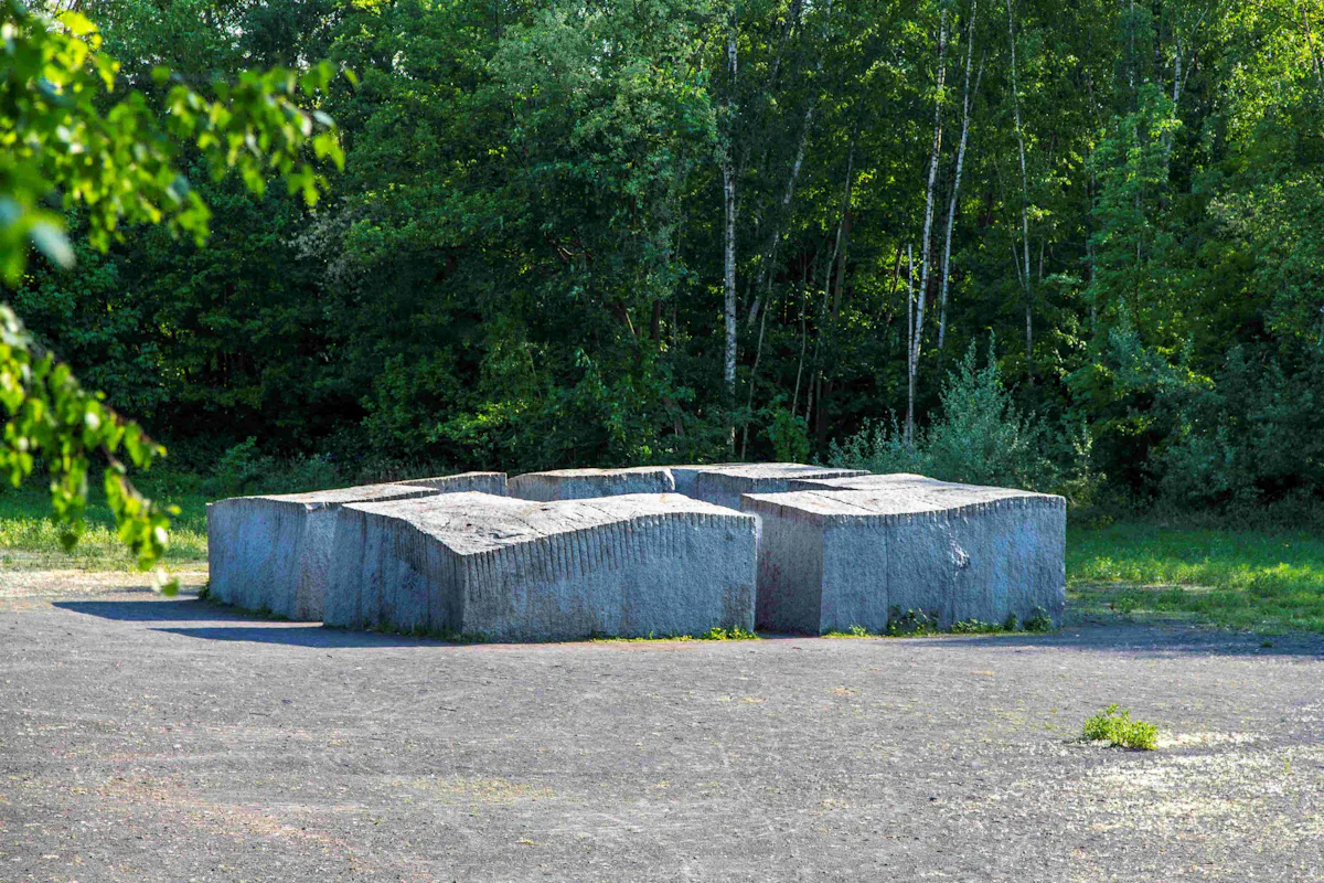Massive, wellenförmig geformte Steinskulptur aus grob bearbeitetem Granit steht auf einer offenen, geschotterten Fläche. Die Skulptur besteht aus mehreren quaderförmigen Elementen mit unregelmäßiger Oberfläche. Im Hintergrund befindet sich ein dichter grüner Laubwald mit verschiedenen Baumarten, darunter Birken. Links im Vordergrund ragen Äste mit grünen Blättern ins Bild.