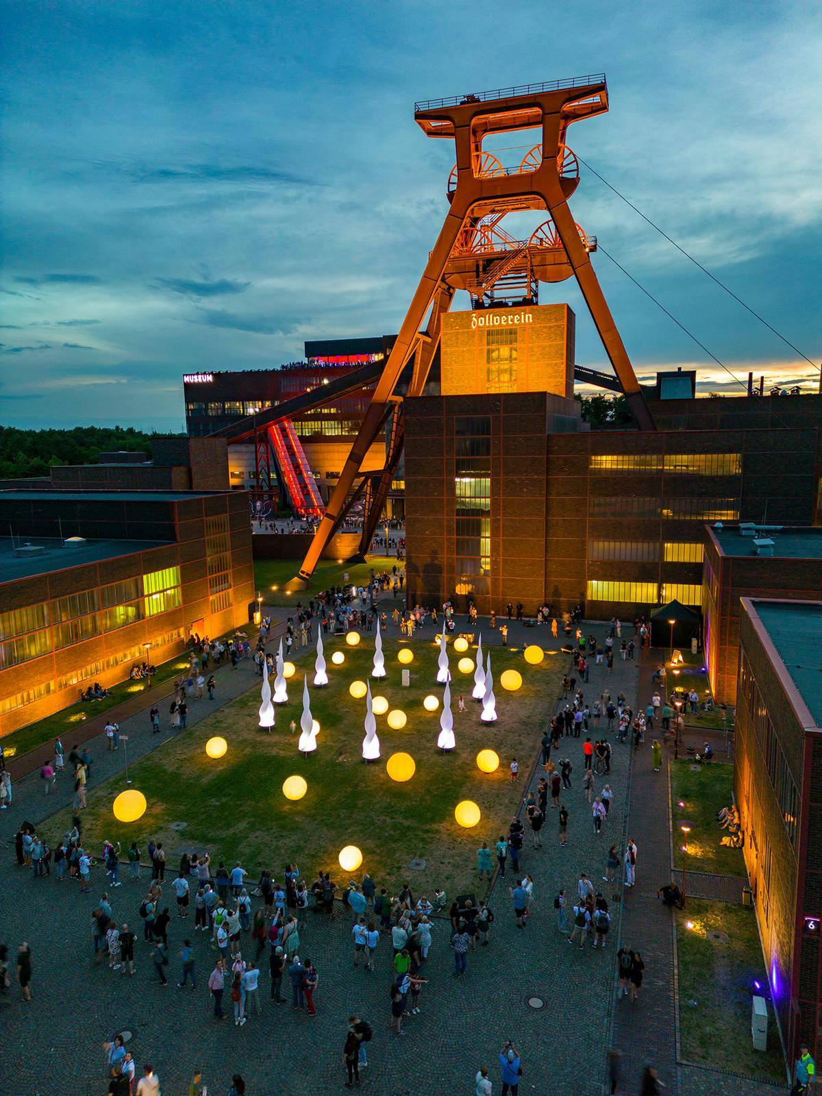 Abendaufnahme des Zollverein-Geländes in Essen mit dem markanten Förderturm im Hintergrund, der orange angestrahlt ist. Auf dem Platz davor sind zahlreiche Menschen versammelt. Die Wiese ist mit leuchtenden gelben Kugeln und weißen, tropfenförmigen Lichtobjekten geschmückt. Die Gebäude rundherum sind ebenfalls in warmem Licht beleuchtet. Die Szene wirkt wie eine Kunst- oder Kulturveranstaltung im Freien.