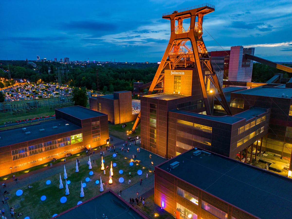 Luftaufnahme des Zollverein-Geländes in Essen während der Dämmerung. Der zentrale Förderturm ist orange beleuchtet und ragt markant über die umgebenden Backsteingebäude. Auf dem Vorplatz befinden sich leuchtende Installationen in Form von weißen Tropfen und farbigen Kreisen. Viele Besucher bewegen sich über das Gelände. Im Hintergrund sind ein voll besetzter Parkplatz und die Stadt mit hell erleuchtetem Horizont zu sehen. Die Atmosphäre wirkt festlich und kulturell.