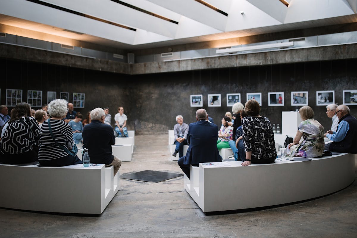 Menschen unterschiedlichen Alters sitzen in einem modernen, halbrund angelegten Raum auf weißen Bänken, die konzentrisch um eine zentrale Fläche angeordnet sind. Sie nehmen an einer Gesprächsrunde oder Diskussion teil. Der Raum hat eine raue Betonoptik mit Oberlichtern an der Decke, die Tageslicht hereinlassen. An den Wänden hängen Fotografien, die wie eine Ausstellung arrangiert sind. Die Atmosphäre wirkt ruhig und konzentriert.