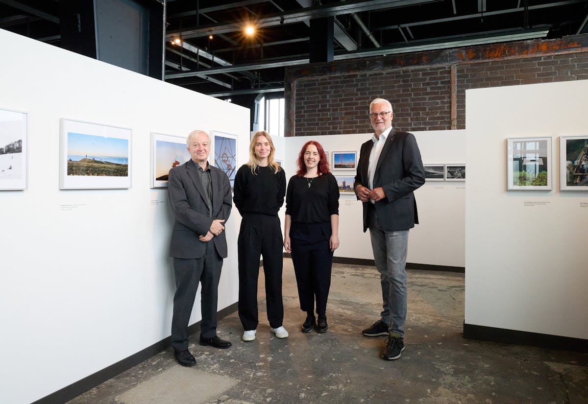 Vier Personen stehen nebeneinander in einer modernen Ausstellungshalle mit weiß gestrichenen Wänden und zahlreichen gerahmten Fotografien. Zwei Männer in Anzügen stehen jeweils außen, dazwischen zwei Frauen in dunkler Kleidung. Alle Personen blicken lächelnd in die Kamera. Der Boden ist aus rauem Beton, die Decke zeigt offene Stahlträger und Beleuchtung. Die Atmosphäre wirkt professionell und freundlich.