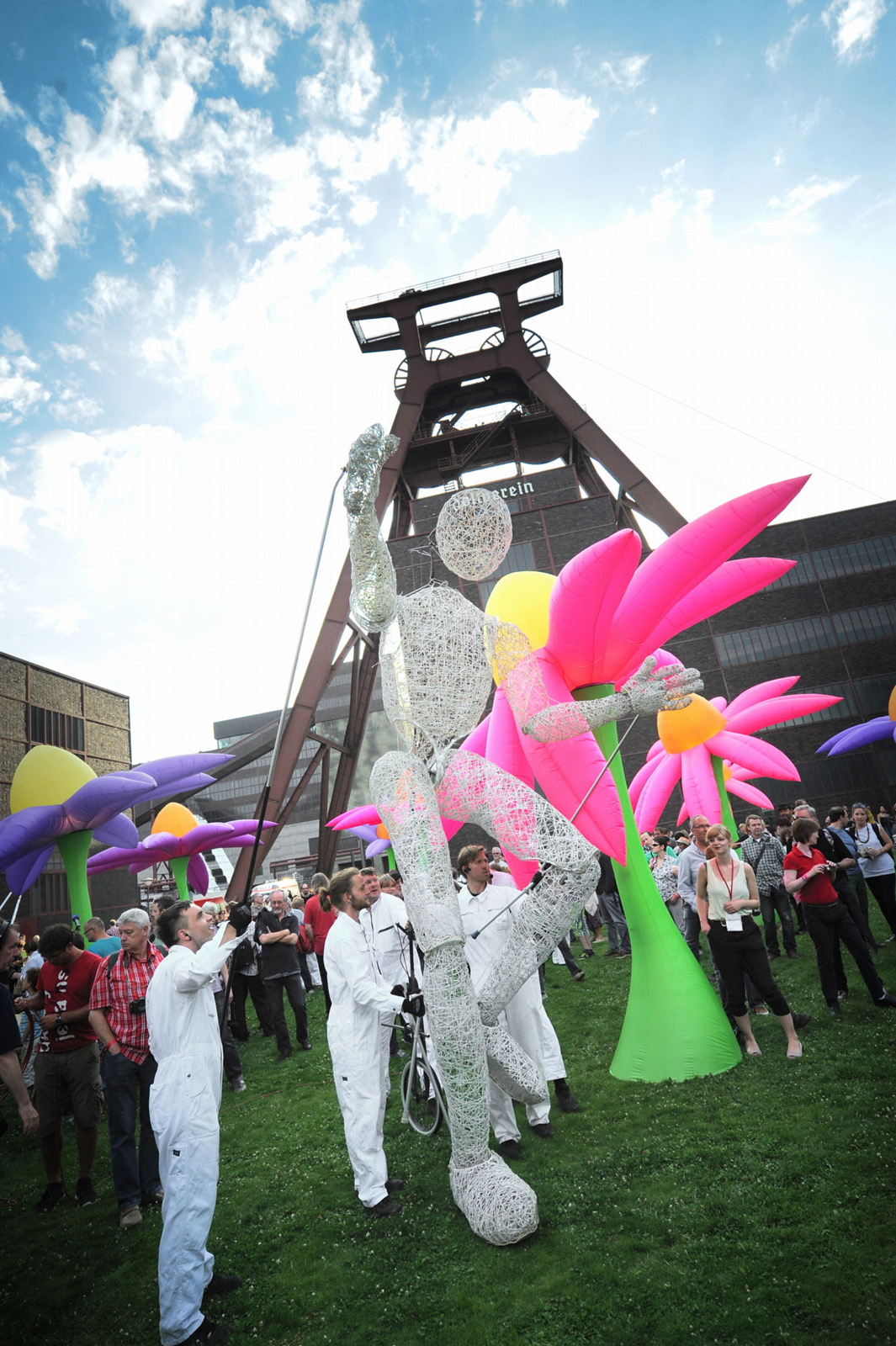 Menschenmenge auf einer Rasenfläche vor dem Förderturm der Zeche Zollverein bei Tageslicht. In der Mitte wird eine große, weiße, drahtartige Puppe von mehreren Personen in weißen Overalls mit Stangen bewegt. Um die Puppe herum stehen übergroße, aufblasbare Blumen in leuchtenden Farben wie Pink, Gelb und Lila. Die Atmosphäre wirkt festlich und fantasievoll, viele Besucher beobachten das Geschehen. Der markante Förderturm ragt im Hintergrund in den blauen, leicht bewölkten Himmel.
