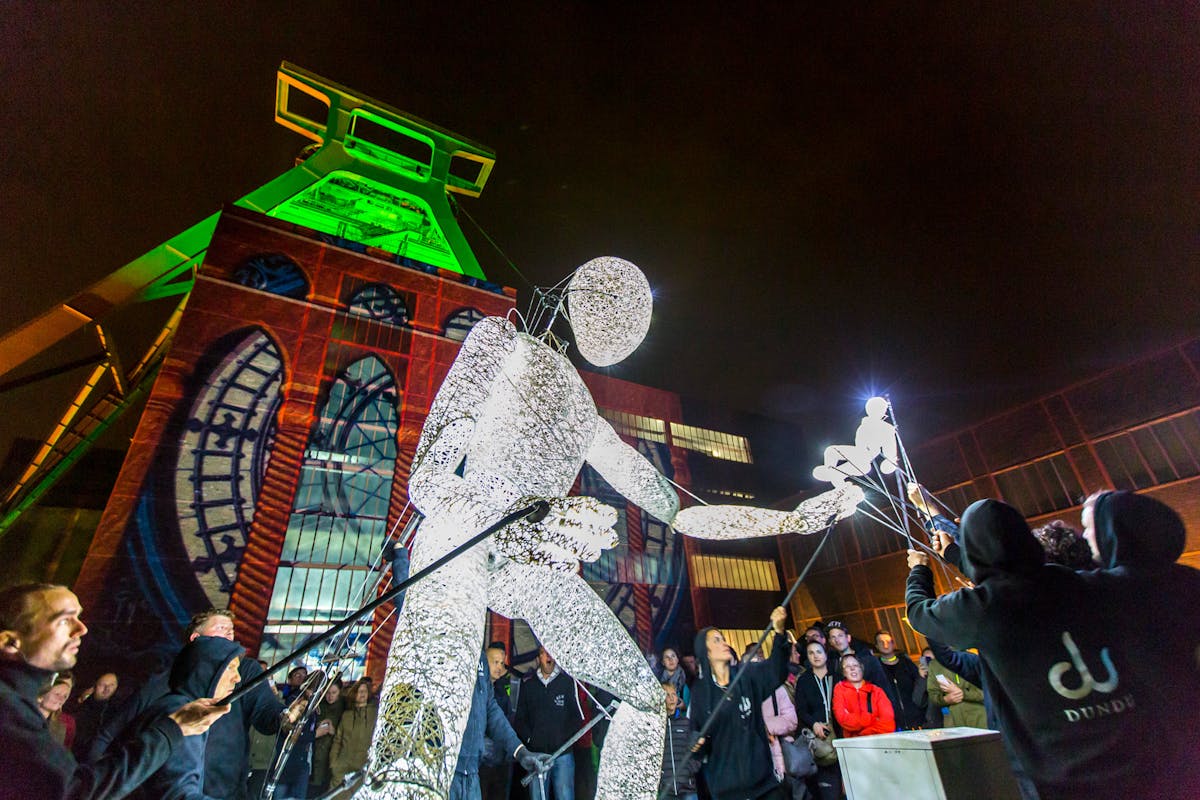Nächtliche Szene auf dem Gelände der Zeche Zollverein mit dem beleuchteten Förderturm im Hintergrund, der grün angestrahlt ist. Im Vordergrund bewegt ein Team aus schwarz gekleideten Personen mit langen Stangen eine große, weiß leuchtende Drahtfigur in menschlicher Form. Die Figur beugt sich leicht nach vorne und streckt einen Arm mit einer kleineren, ebenfalls leuchtenden Figur aus. Im Hintergrund sind viele Zuschauer versammelt, die das Geschehen beobachten. Auf das Gebäude im Hintergrund ist ein buntes Fenster-Motiv projiziert. Die Stimmung ist festlich und künstlerisch.