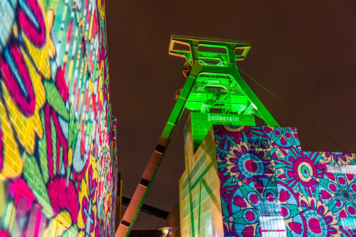 Der Förderturm der Zeche Zollverein ist nachts in intensives grünes Licht getaucht. Auf den angrenzenden Gebäuden werden farbenfrohe, florale und geometrische Muster in kräftigen Tönen wie Pink, Gelb, Blau und Türkis projiziert. Die Lichtinstallation verleiht der Industriekulisse eine lebendige, festliche Atmosphäre. Über einem der Gebäude ist der Schriftzug Zollverein beleuchtet zu sehen. Der Himmel ist dunkel, was die Leuchtkraft der Projektionen zusätzlich betont. Die Szene wirkt wie Teil eines Lichtkunst- oder Kulturfestivals.