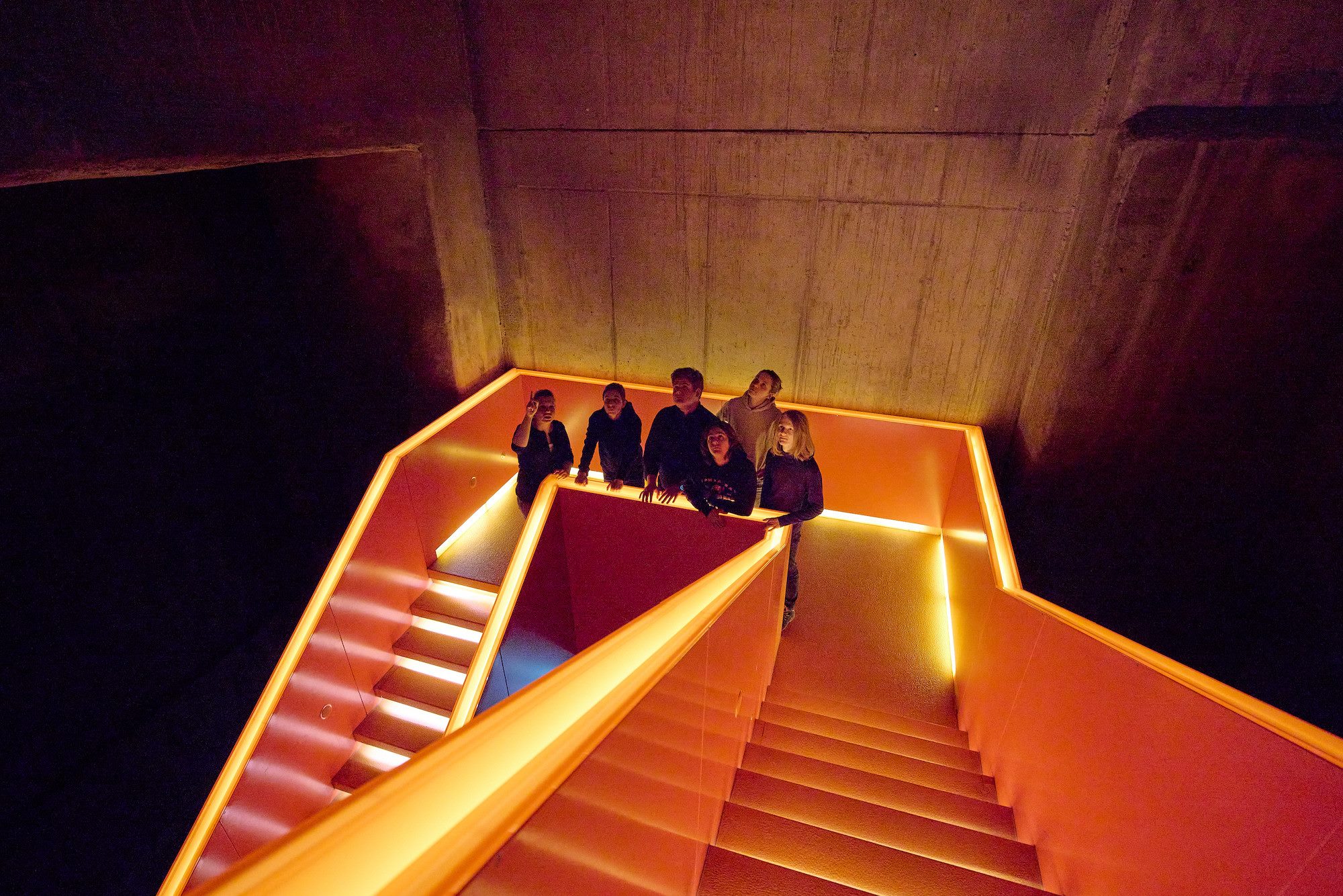 Mehrere Personen stehen auf einer auffällig beleuchteten, orangefarbenen Treppe im Inneren eines Gebäudes mit rohen Betonwänden. Die Stufen und Geländer sind mit indirekter Beleuchtung in warmem Licht hervorgehoben. Die Menschen blicken nach oben, als würden sie eine Führung oder eine besondere Präsentation verfolgen. Die Szenerie wirkt modern und industriell zugleich.