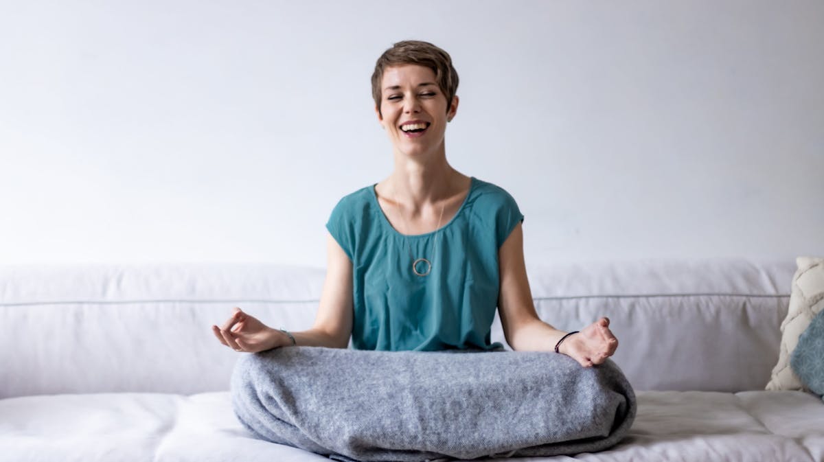 Eine junge Frau mit kurzen braunen Haaren sitzt lächelnd im Lotussitz auf einem hellgrauen Sofa. Sie trägt ein türkisfarbenes Oberteil und hat eine graue Decke über den Beinen. Ihre Hände liegen entspannt auf den Knien mit den Handflächen nach oben, Daumen und Zeigefinger leicht zusammengeführt in einer Meditationshaltung. Im Hintergrund ist eine weiße Wand, rechts liegt ein Kissen mit blauem Muster. Die Stimmung wirkt ruhig und gelassen.