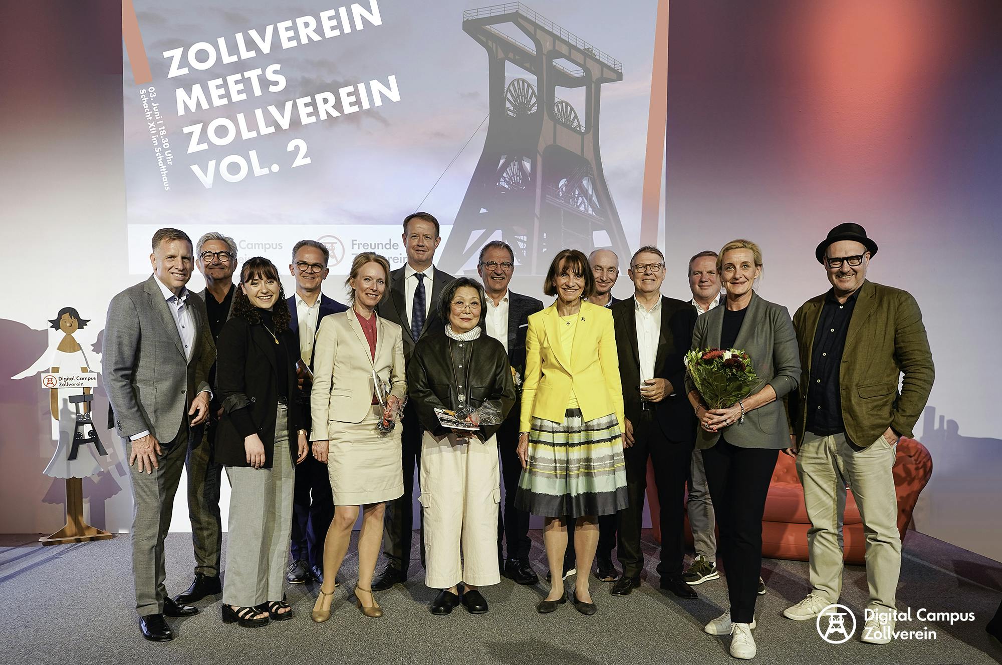 Gruppenfoto von 16 Personen auf einer Bühne vor einer großen Projektionswand mit der Aufschrift Zollverein meets Zollverein Vol. 2. Die Personen stehen eng beieinander, lächeln in die Kamera und wirken festlich gekleidet. Zwei Frauen in der vorderen Reihe halten Blumensträuße und eine weitere Frau hält eine Trophäe in den Händen. Im Hintergrund ist ein stilisiertes Bild des Förderturms der Zeche Zollverein zu sehen. Links neben der Gruppe steht eine Pappfigur mit dem Logo des Digital Campus Zollverein.