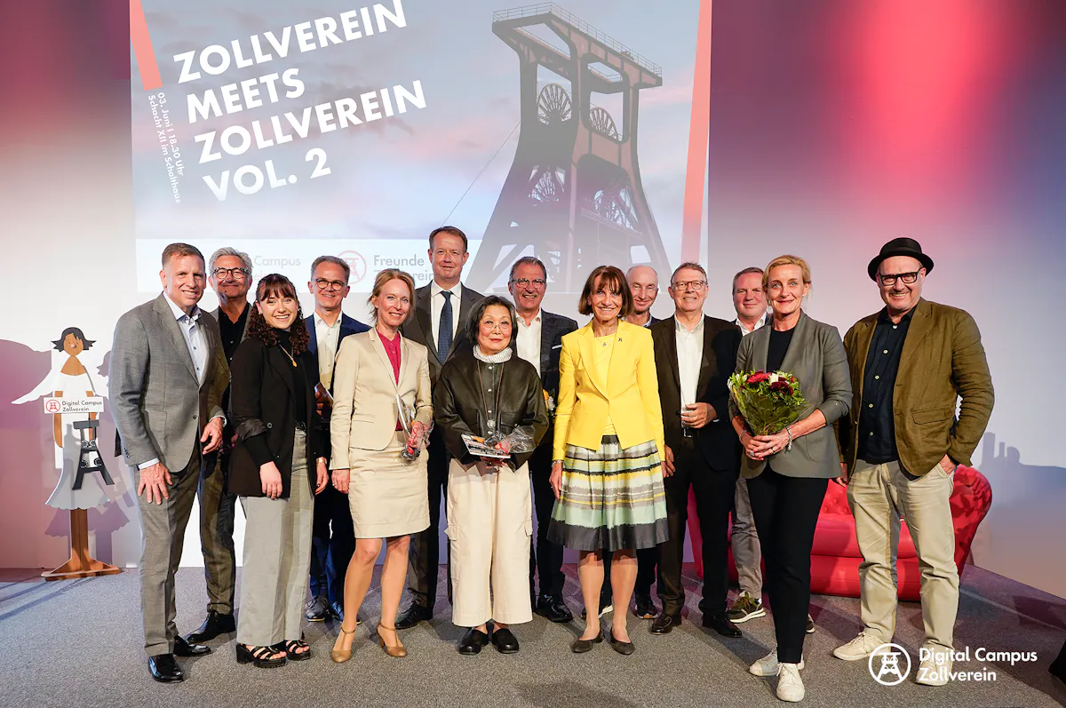 Gruppenfoto von 16 Personen auf einer Bühne vor einer großen Projektionswand mit der Aufschrift Zollverein meets Zollverein Vol. 2. Die Personen stehen eng beieinander, lächeln in die Kamera und wirken festlich gekleidet. Zwei Frauen in der vorderen Reihe halten Blumensträuße und eine weitere Frau hält eine Trophäe in den Händen. Im Hintergrund ist ein stilisiertes Bild des Förderturms der Zeche Zollverein zu sehen. Links neben der Gruppe steht eine Pappfigur mit dem Logo des Digital Campus Zollverein.