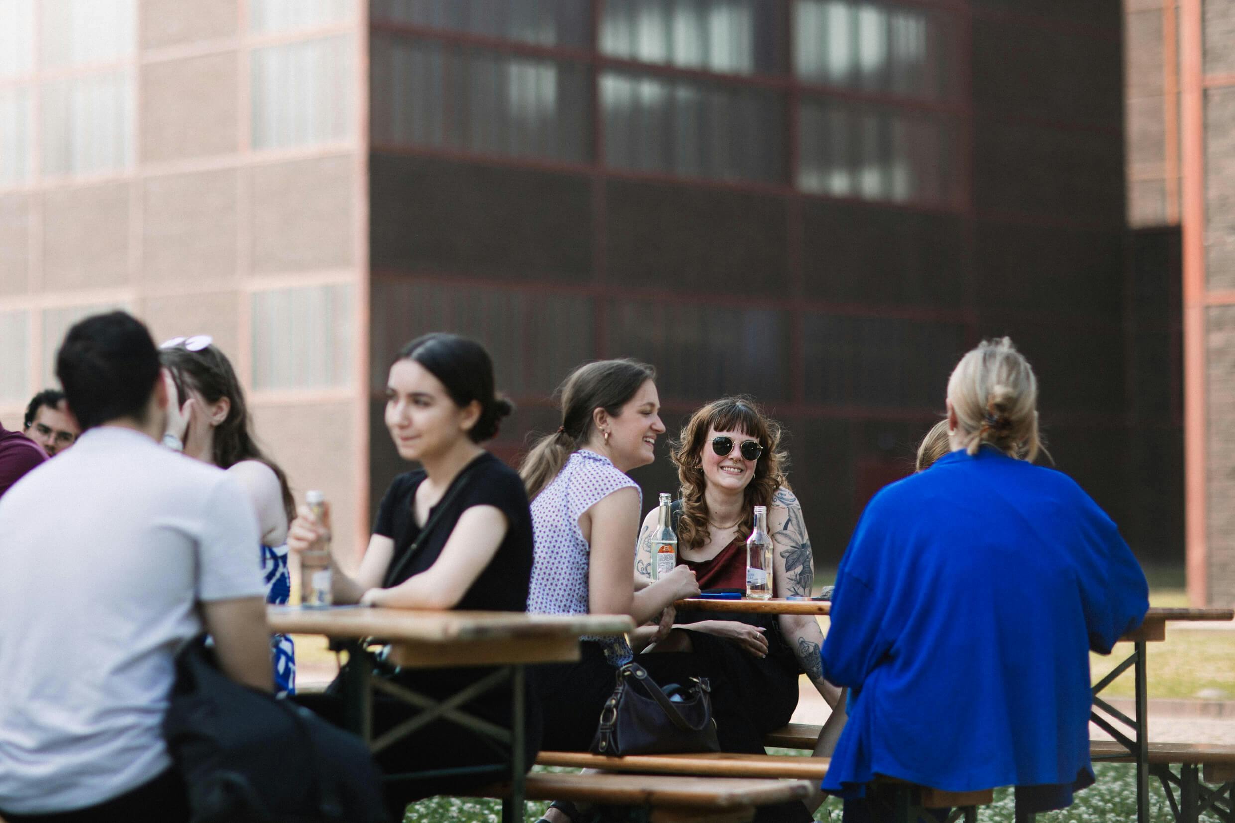 Mehrere junge Erwachsene sitzen an Holzbänken im Freien und unterhalten sich bei Getränken. Die Stimmung ist entspannt und fröhlich. Im Hintergrund sind moderne Ziegelgebäude mit großen Fenstern zu sehen. Die Szene spielt vermutlich im Rahmen einer Veranstaltung oder eines Treffens bei schönem Wetter.