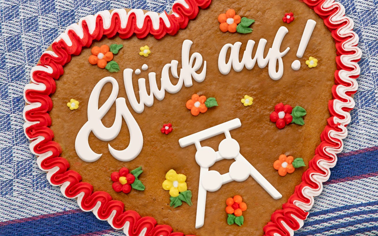 Das Bild zeigt ein Lebkuchenherz auf einem blau-weiß karierten Stoffhintergrund. Das Herz ist mit rotem und weißem Zuckerguss umrandet und trägt in der Mitte die Aufschrift Glück auf in weißer Zuckerschrift. Rund um den Text sind kleine bunte Zuckergussblumen verteilt. Unten rechts auf dem Herz ist ein stilisiertes Fördergerüst aus weißem Zuckerguss abgebildet, das an die Bergbaukultur erinnert. Das Motiv vermittelt einen festlichen, regionalen Bezug, insbesondere zur Tradition des Ruhrgebiets.