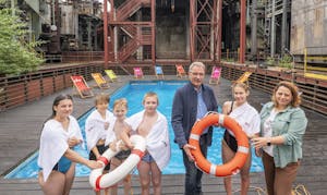 Eine Gruppe von fünf Kindern in Badebekleidung steht mit weißen Handtüchern vor einem kleinen Außenpool auf einem Holzdeck. Zwei Erwachsene, ein Mann und eine Frau, stehen rechts daneben. Der Mann hält einen Rettungsring, die Frau und ein Kind halten einen weiteren. Im Hintergrund befinden sich bunte Liegestühle und industrielle Stahlkonstruktionen mit Treppen und Rohren, die auf ein ehemaliges Industriegelände hindeuten. Die Szene wirkt wie eine Schwimmveranstaltung in ungewöhnlicher Umgebung.