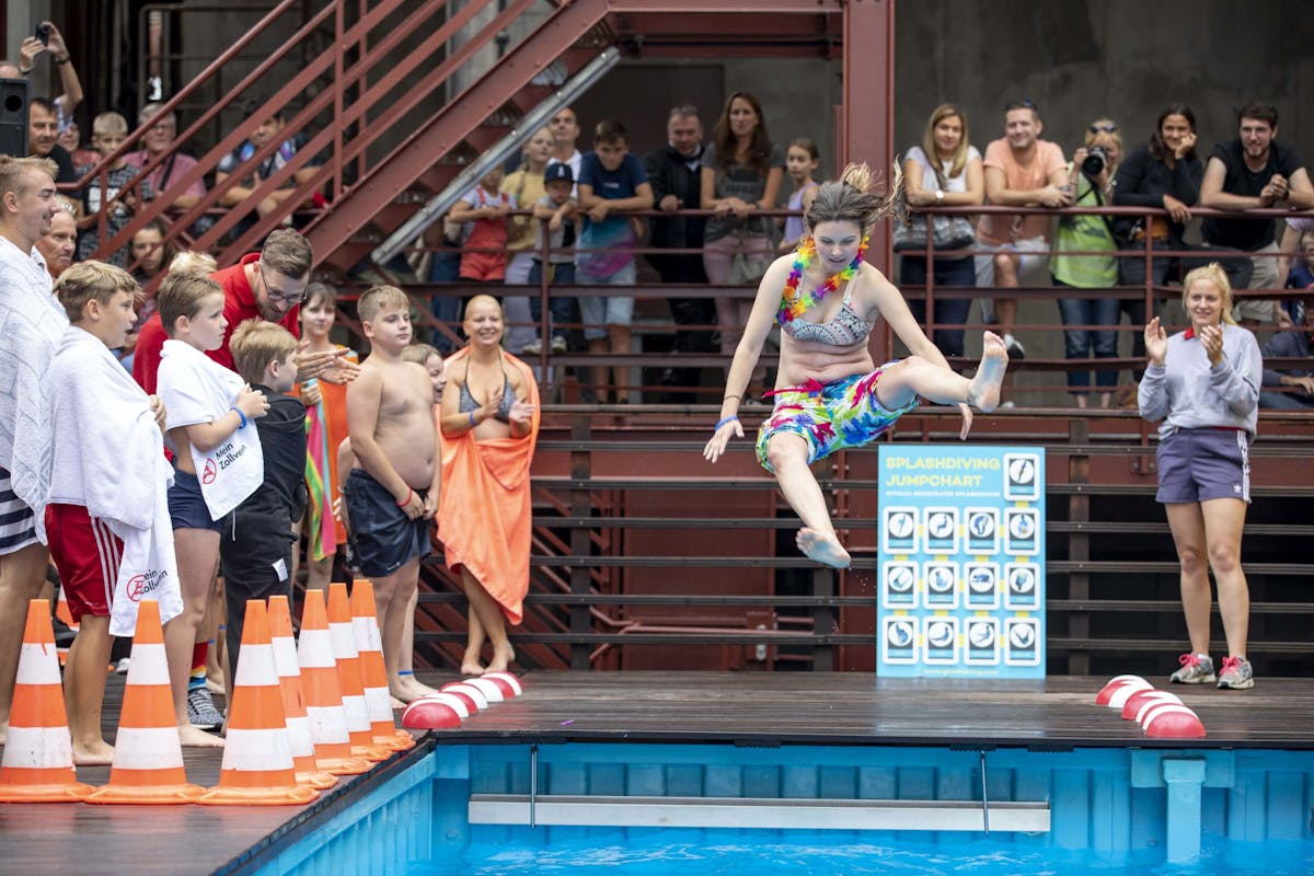Das Bild zeigt eine fröhliche Szene am Beckenrand eines temporären Schwimmbeckens bei einem Splashdiving-Wettbewerb. Im Mittelpunkt springt eine junge Frau in farbenfroher Badebekleidung und Hawaiikette mit angewinkelten Beinen in die Luft, kurz bevor sie ins Wasser eintaucht. Am Rand stehen zahlreiche Kinder und Jugendliche in Badekleidung oder mit Handtüchern, die gespannt zusehen. Im Hintergrund stehen Zuschauer auf einer erhöhten Plattform und schauen neugierig oder machen Fotos. Eine blaue Tafel mit Sprungwertungen und orangene Pylonen markieren den Bereich. Die Stimmung ist lebhaft und sommerlich.
