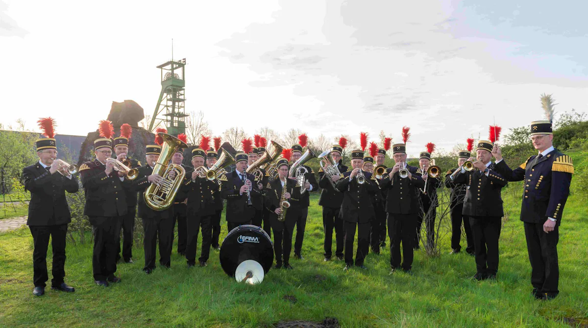 Eine große Blaskapelle spielt im Freien auf einer Wiese vor einem historischen Förderturm. Die Musikerinnen und Musiker tragen traditionelle schwarze Uniformen mit goldenen Knöpfen und hohen Hüten, die mit auffälligen roten Federn geschmückt sind. Die Gruppe besteht überwiegend aus Männern und spielt verschiedene Blechblasinstrumente wie Trompeten, Hörner, Tuben und Posaunen, ergänzt durch ein Saxofon und eine große Trommel mit der Aufschrift Lefima. Ganz rechts steht der Dirigent mit einem weißen Federbusch am Hut und einem Taktstock in der Hand. Die Kulisse zeigt grüne Bäume und Industriearchitektur auf einem ehemaligen Zechengelände.