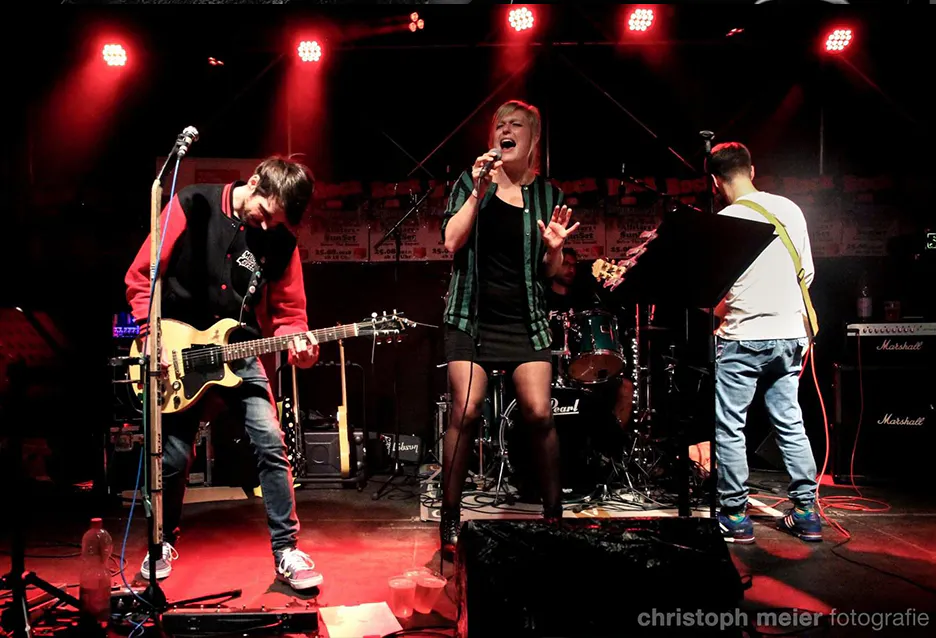 Das Bild zeigt eine Band bei einem Liveauftritt auf einer Bühne unter rotem Scheinwerferlicht. Links spielt ein Gitarrist mit dunklem Haar und roter Jacke eine E-Gitarre, tief nach vorne gebeugt. In der Mitte steht eine Sängerin mit blonder Kurzhaarfrisur, kurzem schwarzen Kleid und kariertem Hemd, die mit ausdrucksstarker Mimik singt. Rechts im Bild steht ein weiterer Musiker mit dem Rücken zur Kamera, spielt Bassgitarre und trägt Jeans sowie ein weißes T-Shirt. Im Hintergrund ist ein Schlagzeuger an einem grünen Drumset zu sehen. Die Bühne ist mit Lautsprechern, Kabeln und Notenständern ausgestattet. Die Stimmung wirkt energiegeladen und intensiv.