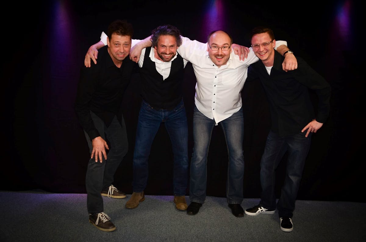Vier Männer stehen nebeneinander vor einem schwarzen Hintergrund mit dezentem lila Licht. Sie beugen sich leicht nach vorne, lachen und haben die Arme freundschaftlich umeinander gelegt. Alle tragen legere Kleidung in dunklen Farben, zwei in weißen Hemden, zwei in schwarzen. Der Boden ist mit grauem Teppich ausgelegt. Die Stimmung wirkt locker und fröhlich, als würden sie zu einer Band oder einem Freundeskreis gehören.