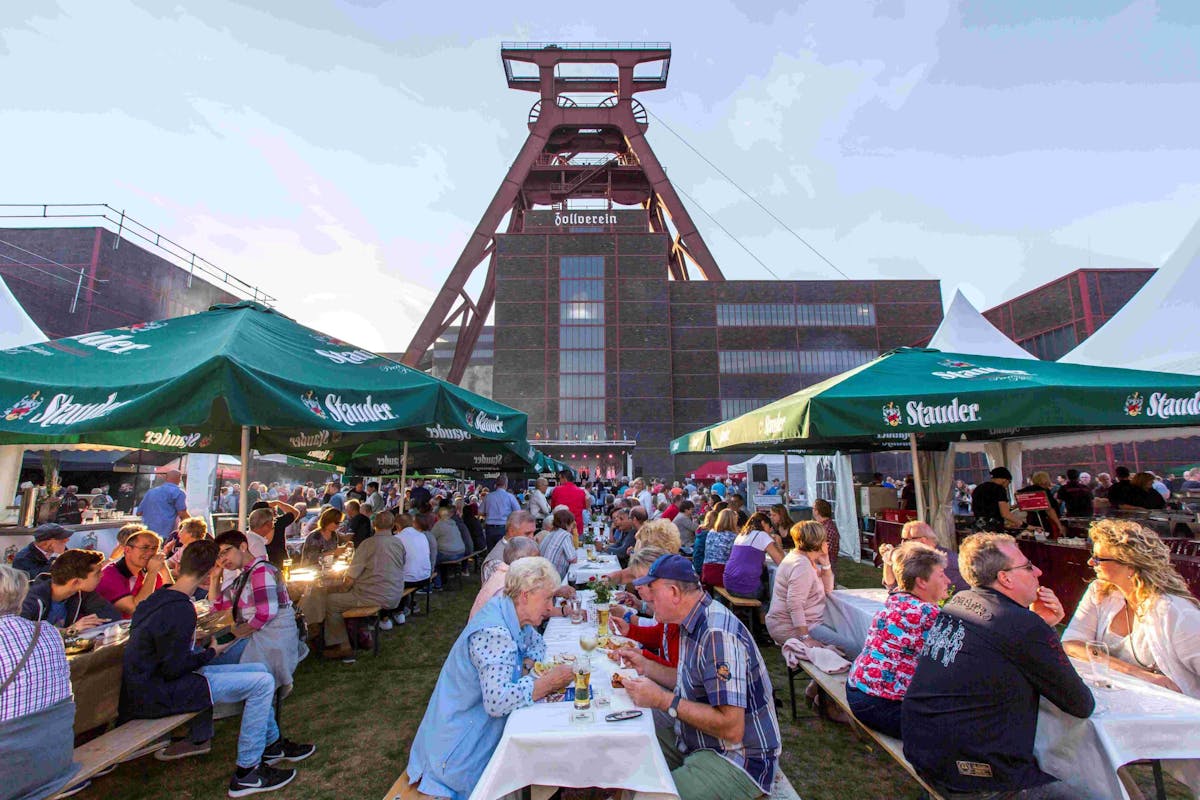 Zahlreiche Menschen sitzen bei sonnigem Wetter unter grünen Sonnenschirmen mit der Aufschrift Stauder an langen Bierbänken und genießen Essen und Getränke. Die Veranstaltung findet vor der Kulisse des bekannten Förderturms der Zeche Zollverein in Essen statt. Im Hintergrund sind weitere Besucher zu sehen, die sich zwischen den Schirmen bewegen. Die Szene vermittelt eine fröhliche, gesellige Atmosphäre bei einem Volksfest.
