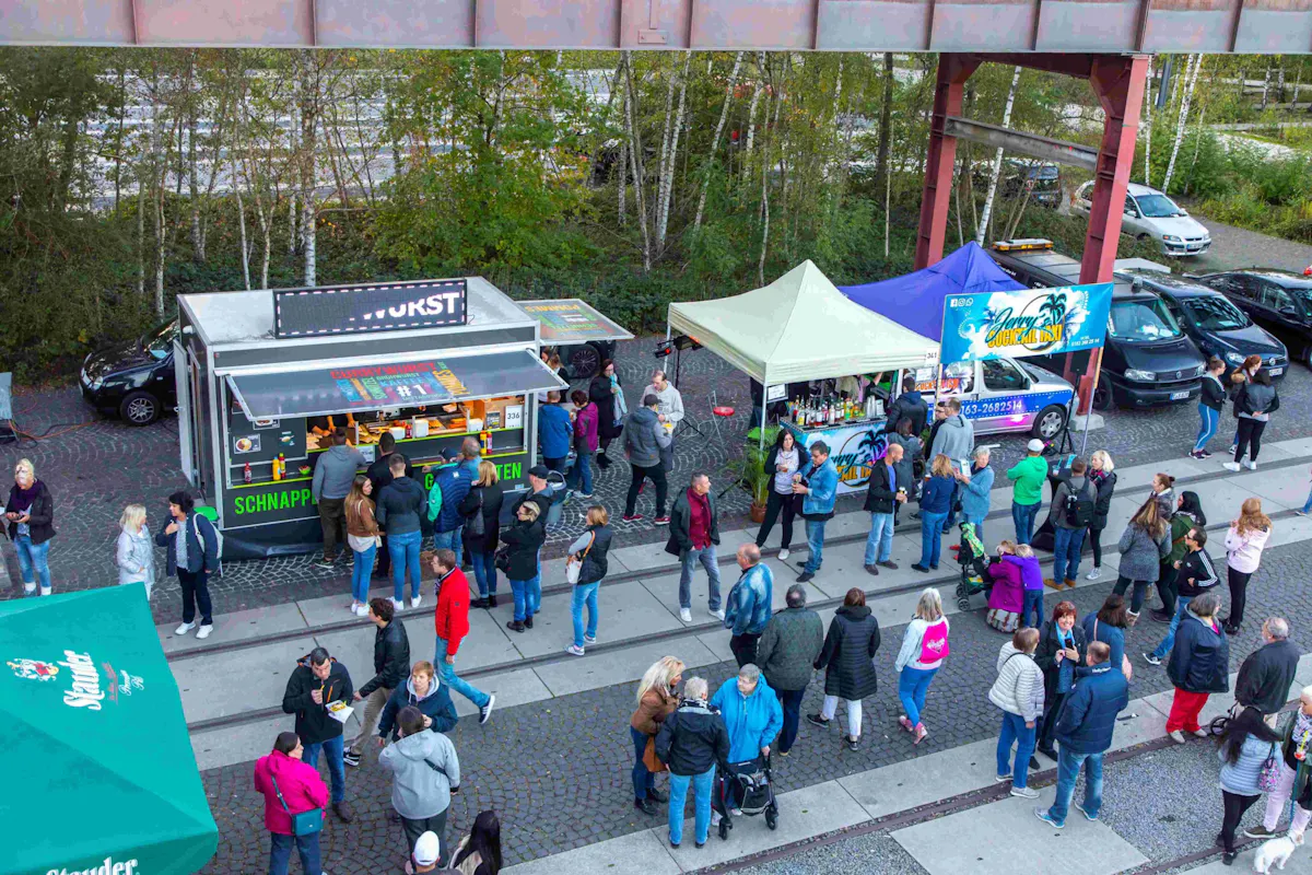 Luftaufnahme eines gut besuchten Straßenfests mit mehreren Foodtrucks und Getränkeständen, vor denen sich viele Menschen in kleinen Gruppen unterhalten oder in Schlangen anstellen. Links steht ein schwarzer Imbisswagen mit der Aufschrift Wurst, daneben ein gelber und ein blauer Cocktailstand mit Palmenbanner. Im Vordergrund sind Besucher jeden Alters unterwegs, einige mit Kinderwagen oder Rollatoren. Der Boden besteht aus Kopfsteinpflaster mit eingelassenen Gleisen, im Hintergrund stehen parkende Autos vor einem Waldstück.