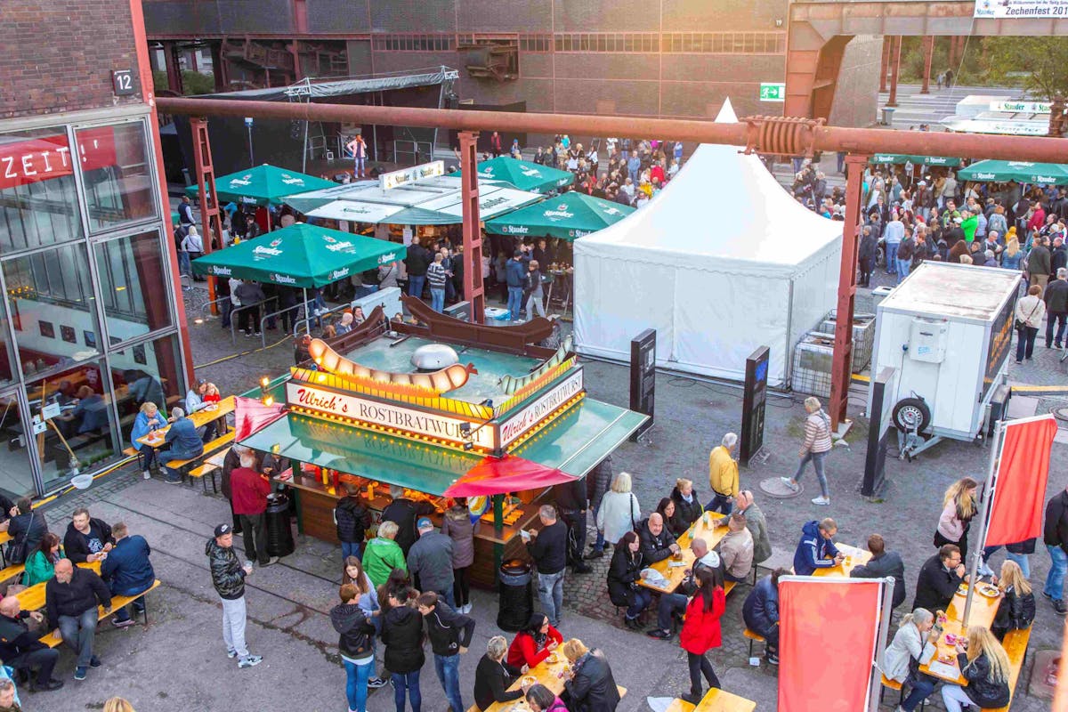 Luftaufnahme eines belebten Festgeländes vor Industriegebäuden mit mehreren Imbissständen und Biergartenbänken. Im Vordergrund steht ein bunter Wurststand mit der Aufschrift Ulrich’s Rostbratwurst, davor und daneben essen viele Menschen an Holztischen. Im Hintergrund sieht man weiße Zelte und grüne Sonnenschirme mit dem Logo der Brauerei Stauder. Dazwischen tummeln sich viele Besucherinnen und Besucher. Links im Bild ein verglastes Gebäude mit weiteren Sitzplätzen im Inneren. Die Atmosphäre ist lebendig und gesellig.
