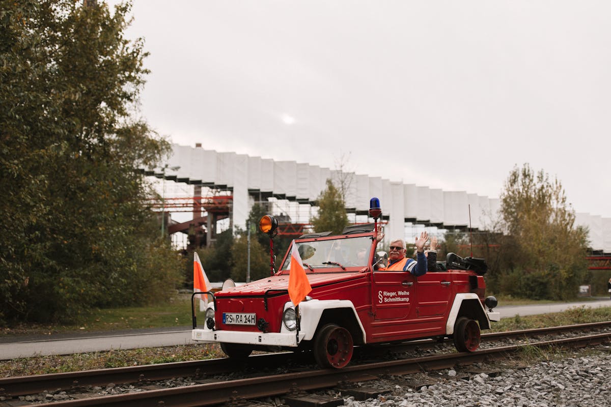 Ein rotes Schienenfahrzeug, umgebaut aus einem offenen Auto mit orangenen Warnflaggen und Blaulicht, fährt auf Gleisen durch eine grüne Umgebung. Zwei ältere Männer in Warnwesten sitzen im Fahrzeug, einer davon winkt freundlich in die Kamera. Im Hintergrund ist ein modernes Industriegebäude mit weißen Fassadenelementen und roten Stahlkonstruktionen zu sehen, umgeben von herbstlich gefärbten Bäumen. Der Himmel ist bewölkt.