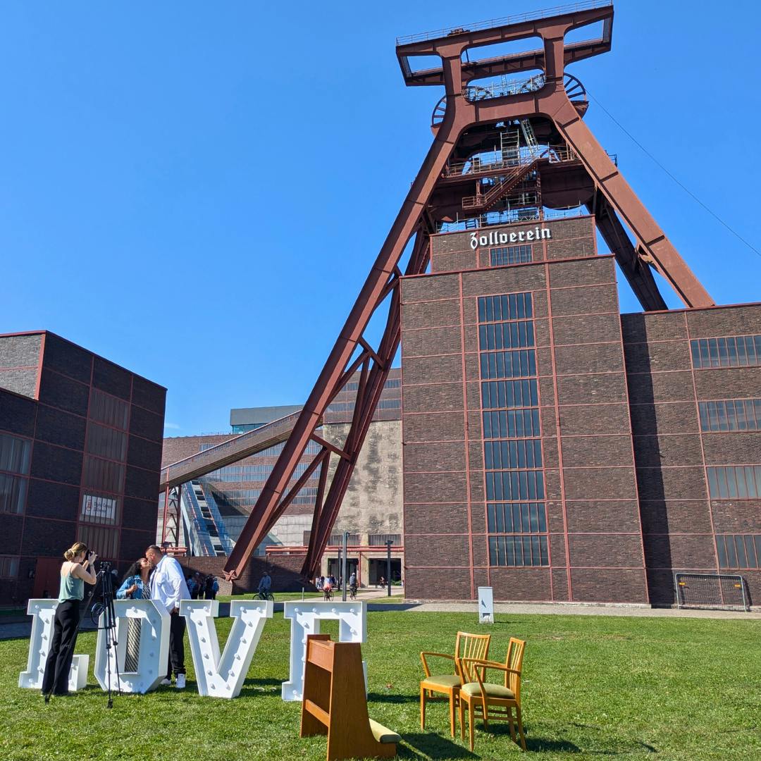 Eine Hochzeitskulisse vor dem markanten Förderturm der Zeche Zollverein bei strahlend blauem Himmel. Im Vordergrund stehen große weiße Buchstaben, die das Wort LOVE formen. Ein Brautpaar küsst sich daneben, während eine Fotografin den Moment festhält. Auf der Wiese befinden sich auch einige gelbe Stühle und ein kleines Rednerpult.