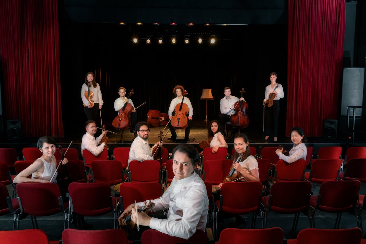 Eine Gruppe von Musikerinnen und Musikern mit Streichinstrumenten posiert in einem Theater oder Konzertsaal. Einige sitzen in den vorderen Reihen mit ihren Instrumenten in der Hand, andere stehen auf der Bühne mit Violinen, Bratschen und Celli. Alle tragen weiße Hemden oder Blusen und schauen freundlich in Richtung Kamera. Der Saal ist mit roten Theatersesseln und roten Vorhängen ausgestattet. Über der Bühne hängen mehrere warme Scheinwerfer. Die Szene wirkt wie ein Gruppenporträt eines Kammerorchesters oder eines Ensembles vor einem Auftritt oder zur Promotion.