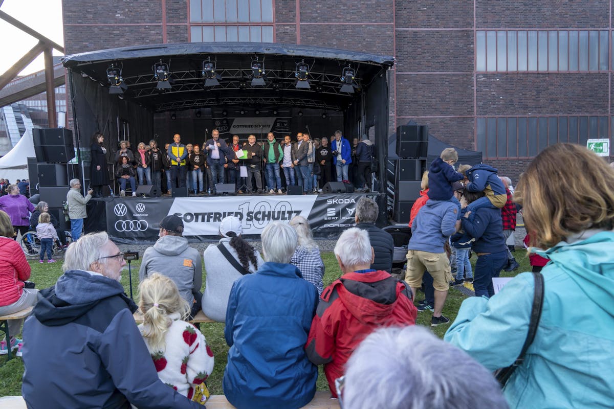 Eine große Gruppe von Menschen steht auf einer Bühne im Freien vor einem Industriegebäude. In der Mitte spricht ein Mann mit Mikrofon, daneben halten Personen Unterlagen in der Hand. Vor der Bühne sitzen und stehen zahlreiche Zuschauerinnen und Zuschauer, darunter Kinder, ältere Menschen und Familien. Die Bühne ist mit einem Banner geschmückt, auf dem unter anderem die Logos von Volkswagen, Audi und weiteren Automarken sowie der Schriftzug Gottfried Schultz zu sehen sind. Einige Kinder sitzen auf den Schultern der Erwachsenen, um besser sehen zu können.