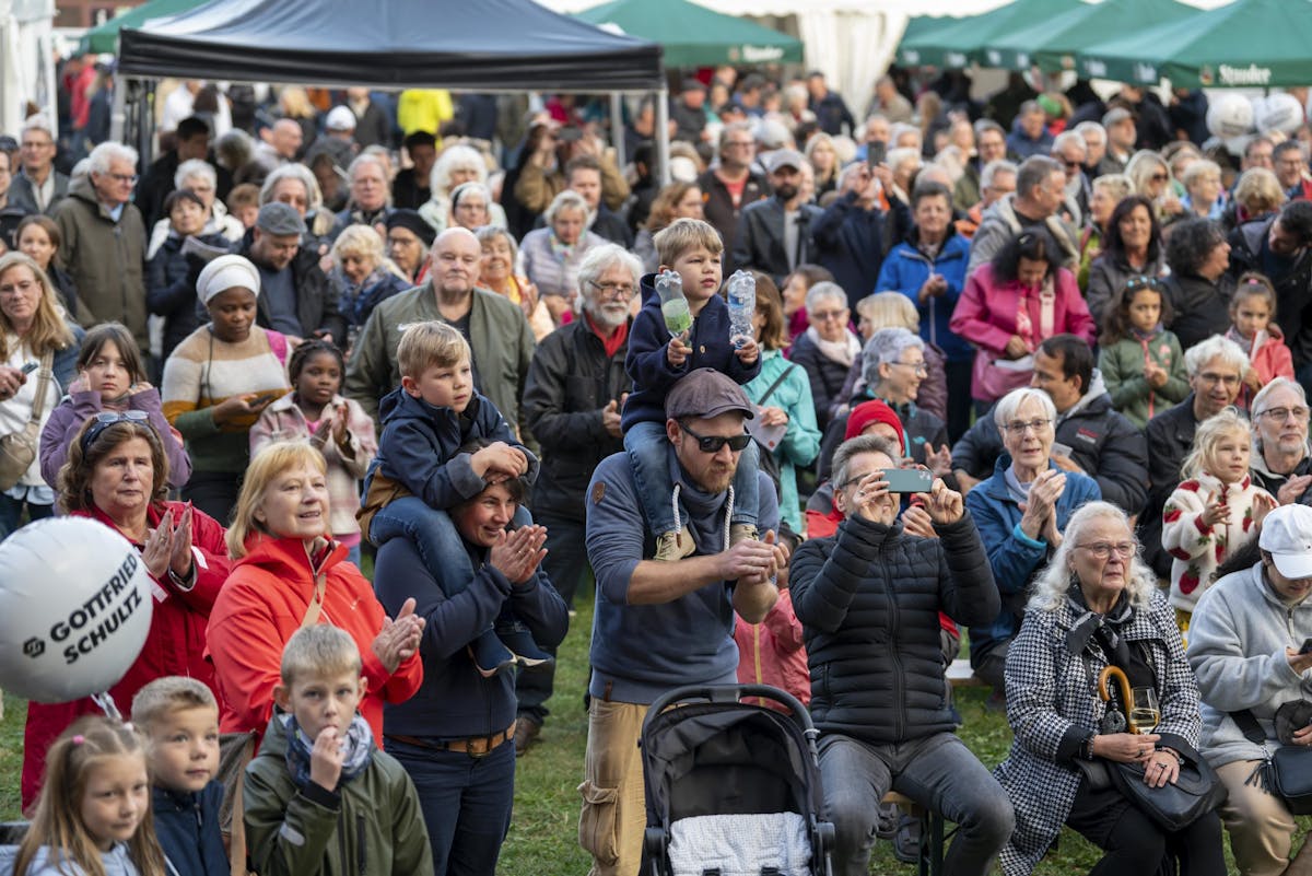 Eine große Menschenmenge unterschiedlichen Alters und Herkunft steht dicht gedrängt auf einem Festgelände. Im Vordergrund sind Kinder, Eltern und ältere Menschen zu sehen, einige mit Kinderwagen oder Luftballons. Zwei Jungen sitzen auf den Schultern von Erwachsenen, einer hält zwei bunte Plastikflaschen in den Händen. Viele Menschen klatschen, lächeln oder filmen mit dem Handy. Im Hintergrund stehen grüne Pavillons und weiße Zelte, die vermutlich zu Verkaufsständen oder Informationsständen gehören. Die Stimmung wirkt fröhlich und aufmerksam.