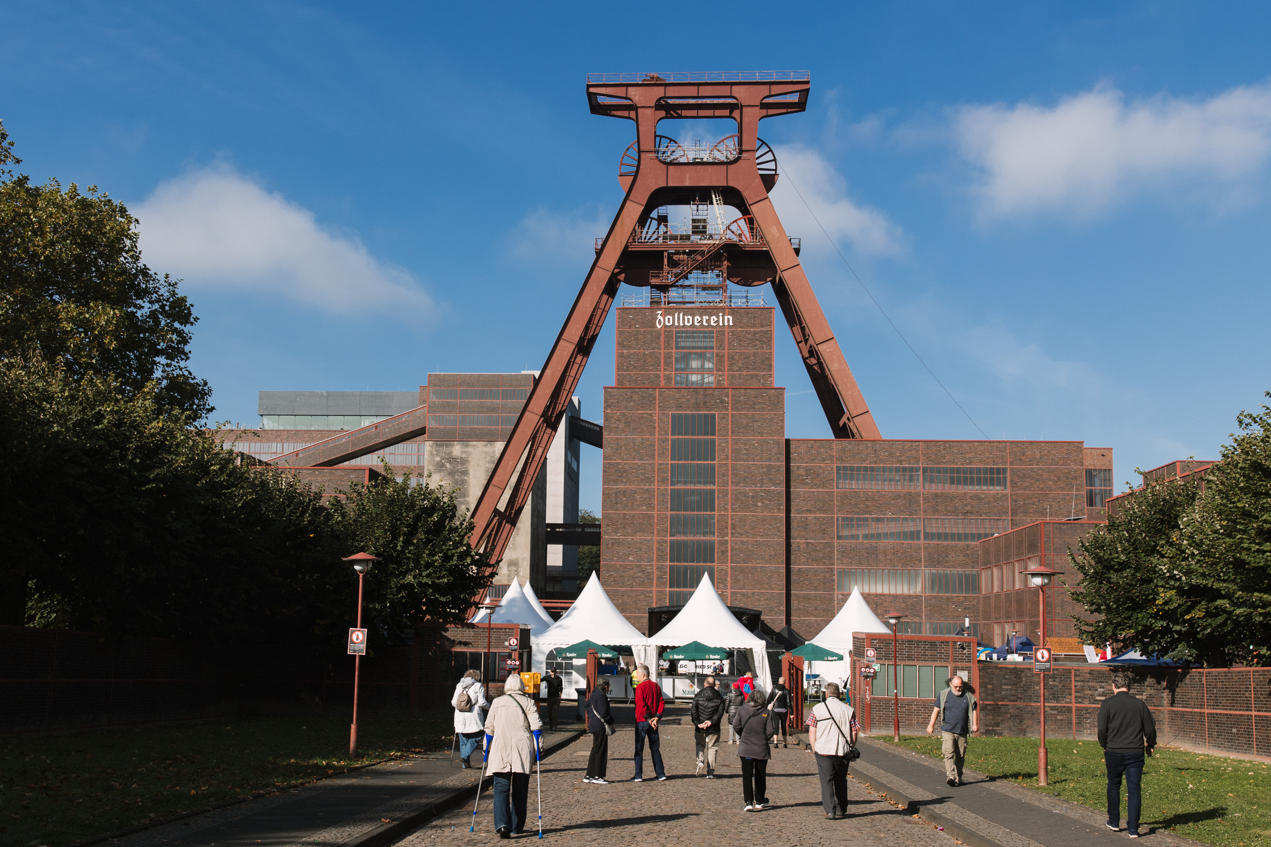 Mehrere Menschen gehen bei sonnigem Wetter auf das Gelände der Zeche Zollverein in Essen zu, wo weiße Festzelte unter dem markanten Förderturm mit der Aufschrift Zollverein aufgebaut sind. Das imposante Backsteingebäude und der rote Stahlförderturm ragen vor einem blauen Himmel mit wenigen Wolken in die Höhe. Links und rechts des Weges stehen Bäume und Laternen, das Gelände wirkt gepflegt und einladend.