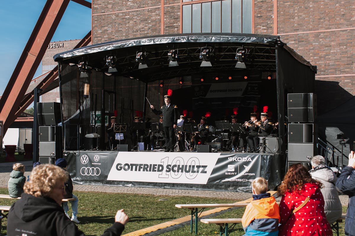 Eine Blaskapelle in traditioneller Bergmannsuniform mit schwarzen Jacken und roten Federbüschen spielt auf einer Bühne vor der Kulisse der Zeche Zollverein. In der Mitte steht der Dirigent mit erhobenem Taktstock. Vor der Bühne sitzen und stehen Zuschauer auf Bierbänken, einige applaudieren. Ein großes Banner an der Bühne zeigt die Aufschrift Gottfried Schultz 100 sowie verschiedene Automarken. Die Sonne scheint und das Wetter ist klar.