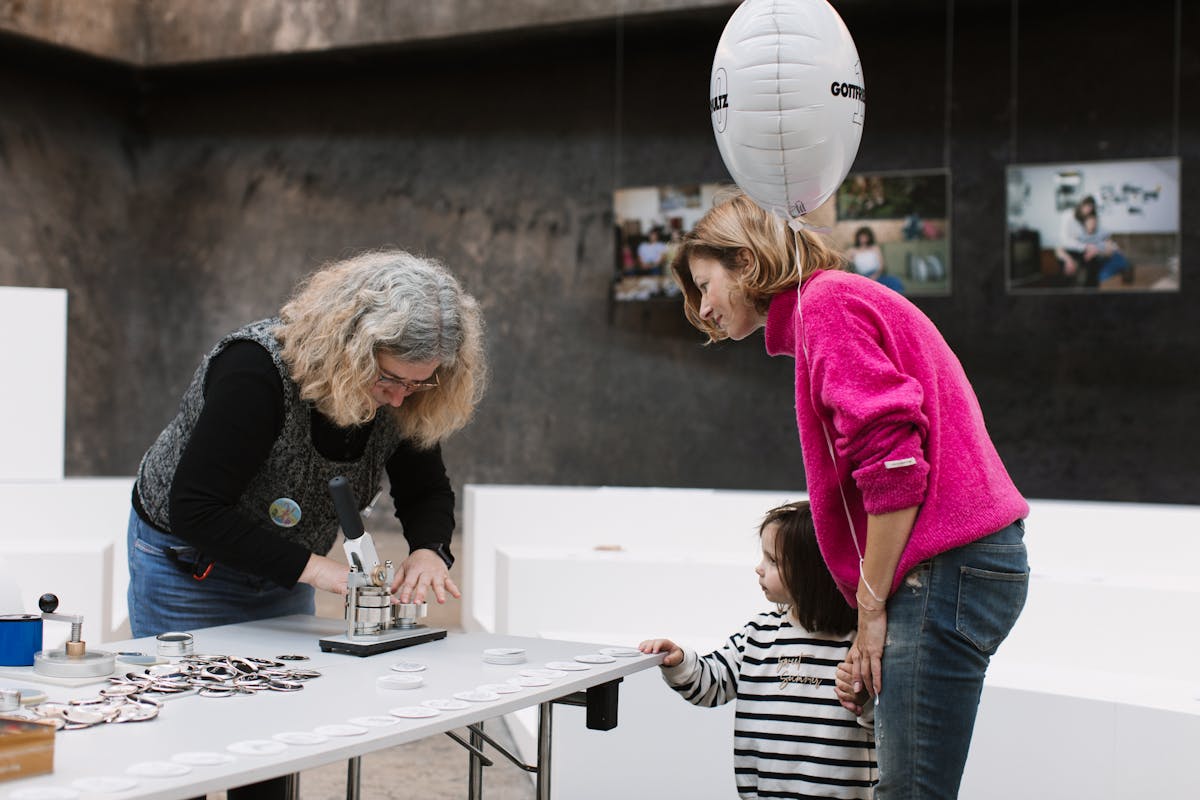 Eine Frau mit grauen, lockigen Haaren presst mit einer Maschine Buttons auf einem Tisch. Auf dem Tisch liegen zahlreiche vorbereitete Buttonteile. Eine andere Frau in pinkem Pullover steht daneben und beobachtet den Vorgang gemeinsam mit einem kleinen Kind im gestreiften Pullover, das ihre Hand hält. Das Kind schaut neugierig auf die Maschine. Die Frau trägt einen weißen Ballon mit der Aufschrift Gottfried Schultz. Im Hintergrund hängen Fotos an einer dunklen Wand.