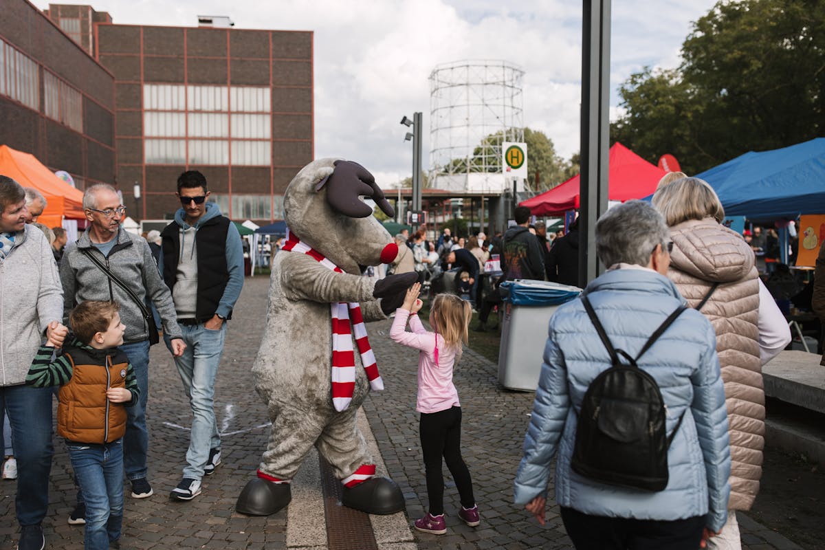 Auf einem belebten Festgelände gibt ein als Rentier verkleidetes Maskottchen einem kleinen Mädchen ein High-Five. Das Rentierkostüm ist grau mit roter Nase, weißen Hörnern und einem rot-weiß gestreiften Schal. Um die beiden herum gehen Erwachsene und Kinder zwischen bunten Marktständen mit Zeltdächern umher. Im Hintergrund sind ein altes Industriegebäude und ein Gasometer zu sehen. Die Atmosphäre ist lebendig und freundlich.