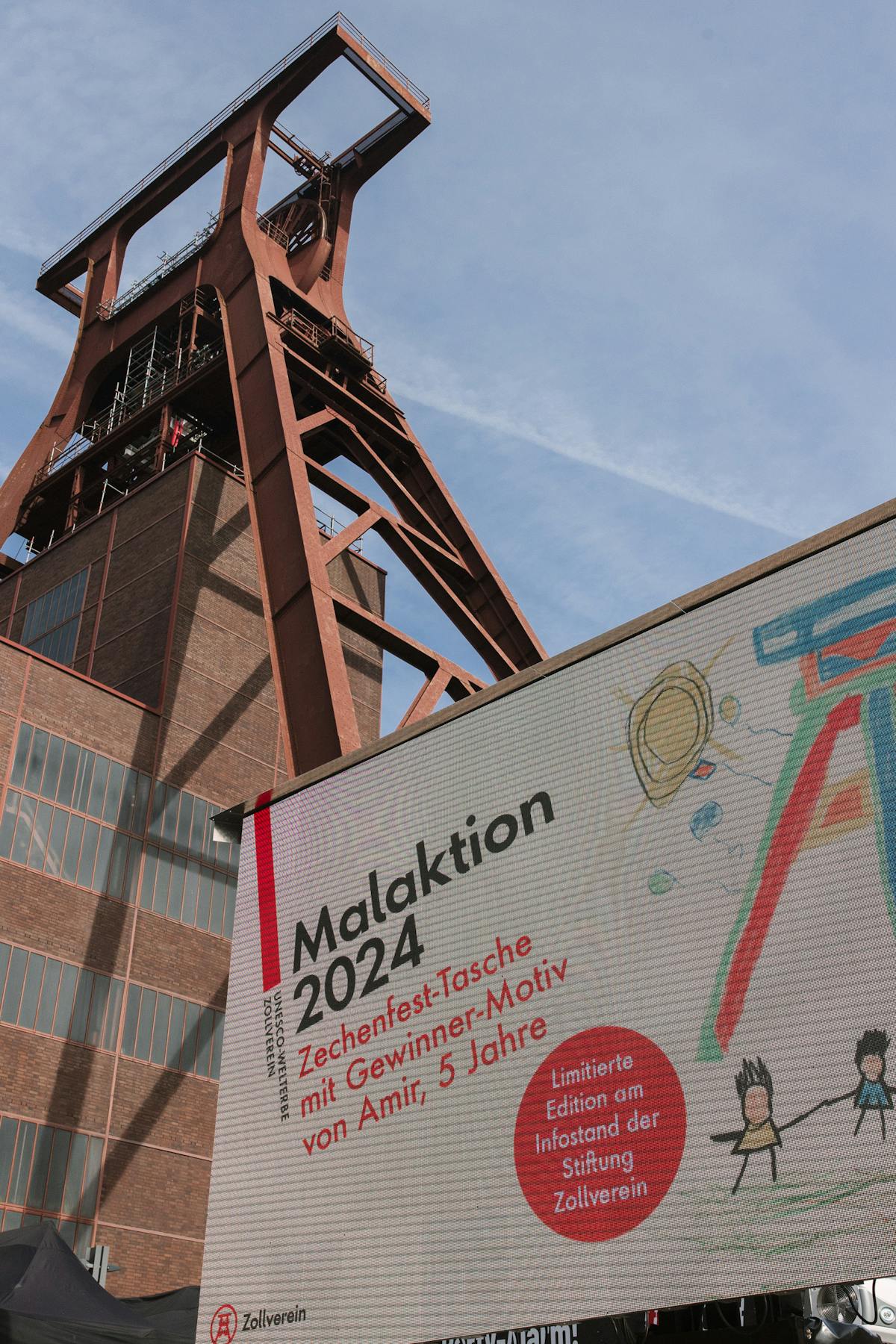 Im Vordergrund ist ein großes Werbebanner zur Malaktion 2024 des Zechenfests Zollverein zu sehen. Darauf steht, dass eine Zechenfest-Tasche mit dem Gewinner-Motiv von Amir, 5 Jahre alt, als limitierte Edition am Infostand der Stiftung Zollverein erhältlich ist. Rechts auf dem Banner ist eine bunte Kinderzeichnung abgebildet, darunter zwei Strichmännchen, die sich an den Händen halten. Im Hintergrund ragt der markante Förderturm der Zeche Zollverein in den Himmel.