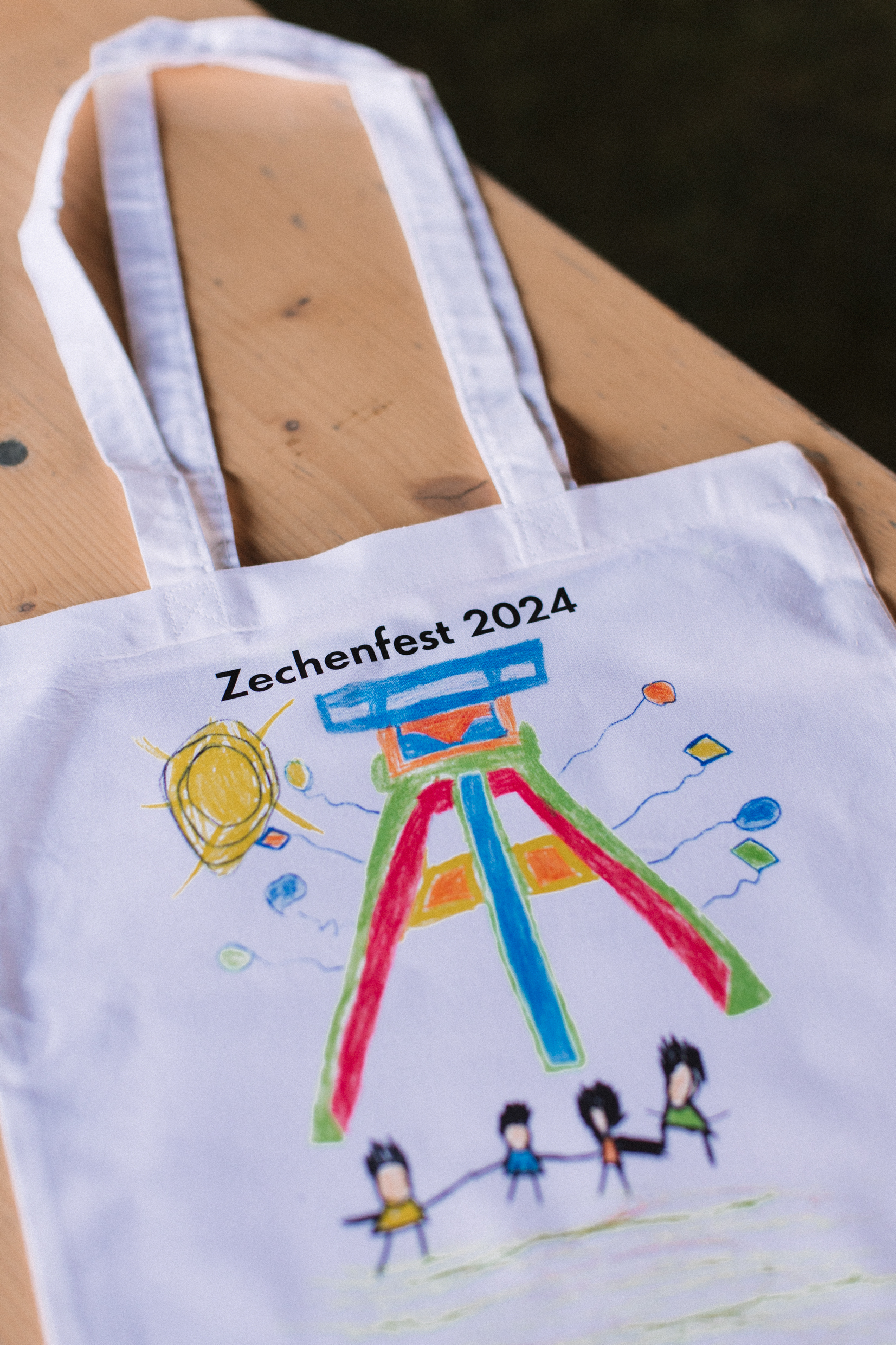 Eine weiße Stofftasche liegt auf einem Holztisch. Auf der Tasche steht in schwarzer Schrift Zechenfest 2024. Darunter ist eine bunte Kinderzeichnung abgebildet, die das Fördergerüst der Zeche Zollverein in leuchtenden Farben zeigt. Um das Gerüst sind Luftballons und eine große gelbe Sonne gemalt. Unten auf der Zeichnung halten sich vier fröhliche Strichmännchen an den Händen.