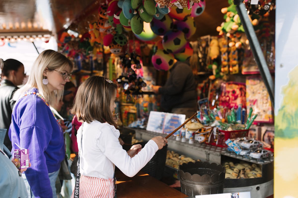 Ein junges Mädchen steht an einem bunten Jahrmarktstand und hält eine Angelrute in der Hand, mit der sie versucht, ein kleines Spielzeug zu angeln. Neben ihr steht eine blonde Frau in lila Pullover, vermutlich ihre Mutter, die ihr zusieht. Über ihnen hängen bunte Plüschtiere und rund um den Stand sind zahlreiche Spielwaren und Preise ausgestellt. Die Szene spielt sich bei Tageslicht in fröhlicher Atmosphäre ab.
