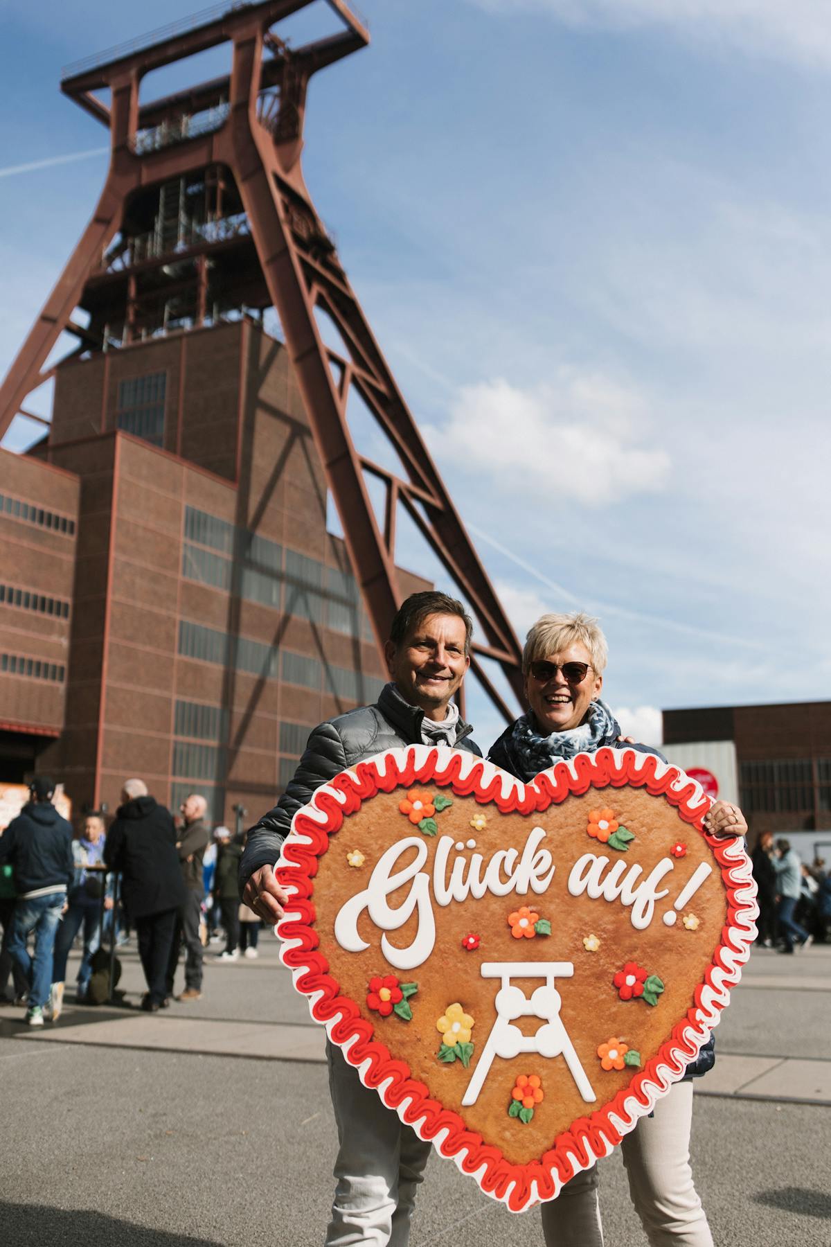 Ein lächelndes Paar steht vor dem markanten Förderturm der Zeche Zollverein und hält gemeinsam ein überdimensionales Lebkuchenherz in den Händen. Das Herz ist mit rotem Zuckerguss verziert und trägt die weiße Aufschrift Glück auf sowie bunte Blumen und ein stilisiertes Fördergerüst. Um das Paar herum befinden sich weitere Besucherinnen und Besucher des Festgeländes. Der Himmel ist blau mit vereinzelten Wolken.