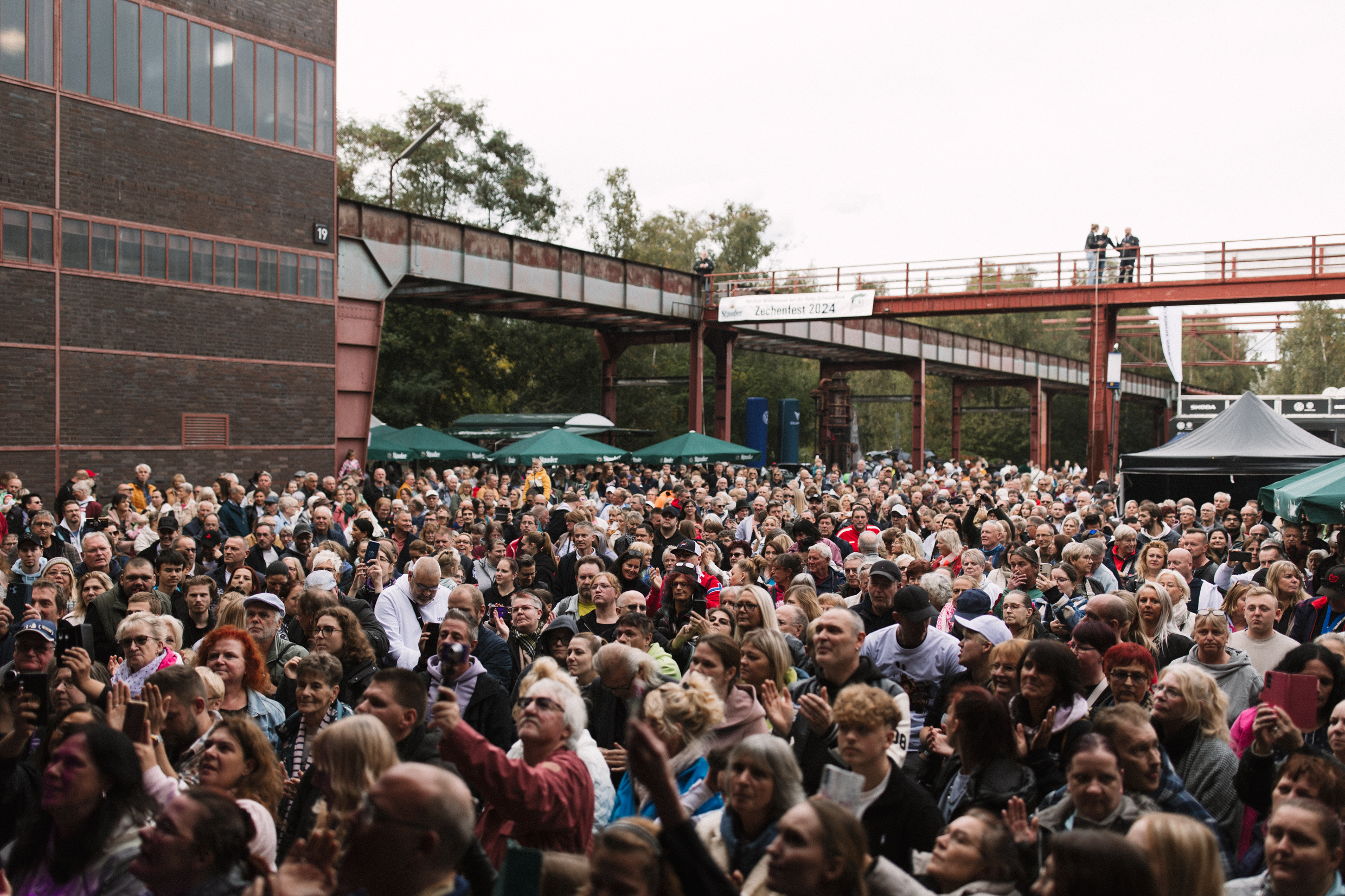 Eine dichte Menschenmenge steht eng beisammen auf dem Gelände der Zeche Zollverein beim Zechenfest 2024. Die Menschen wirken fröhlich und aufmerksam, viele halten Smartphones in der Hand oder klatschen. Im Hintergrund sind grüne Pavillons, ein schwarzes Festzelt und ein roter Steg mit Bannern und Besuchern zu sehen. Die Szenerie ist umgeben von alten Industriebauten mit roten Stahlträgern und Backsteinfassaden.