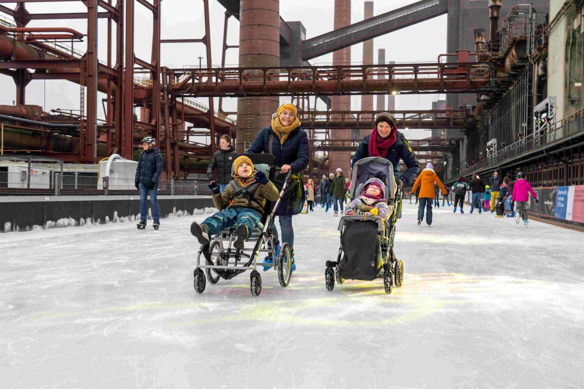 Menschen genießen eine winterliche Aktivität auf einer großen Eisbahn im Freien, die sich in einem ehemaligen Industriekomplex mit rostfarbenen Stahlkonstruktionen und Schornsteinen befindet. Im Vordergrund schieben zwei Frauen jeweils ein Kind im Rollstuhl beziehungsweise im Buggy über das Eis. Die Szene zeigt ein inklusives und fröhliches Miteinander. Weitere Personen im Hintergrund fahren Schlittschuh oder spazieren auf dem Eis, eingepackt in warme Winterkleidung. Die Atmosphäre ist lebendig und herzlich.